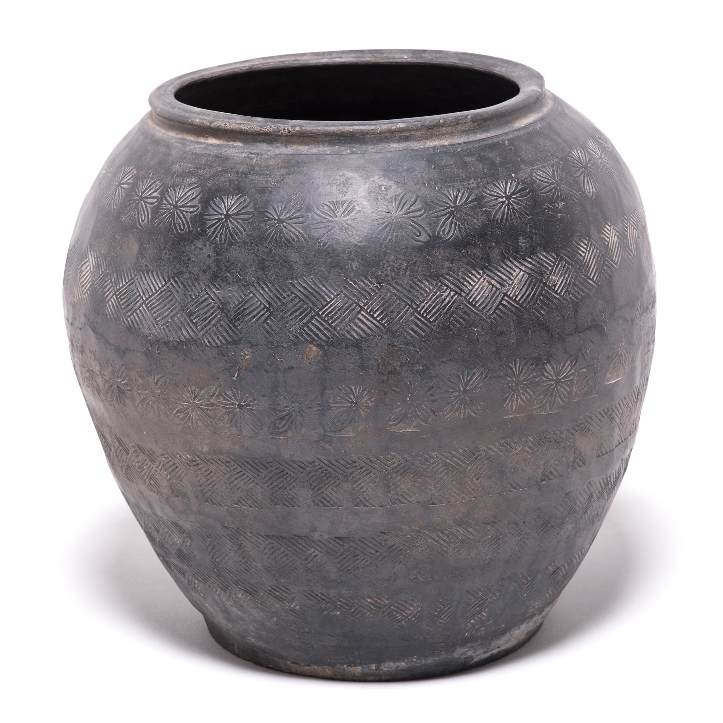 Over a century ago, a gifted potter from northern China crafted this organic clay jar out of rich dark river clay mined from the river basins of Asia. The surface is stamped bands of symbols: hashmarks, flowers, and cross-checking. The effect is a