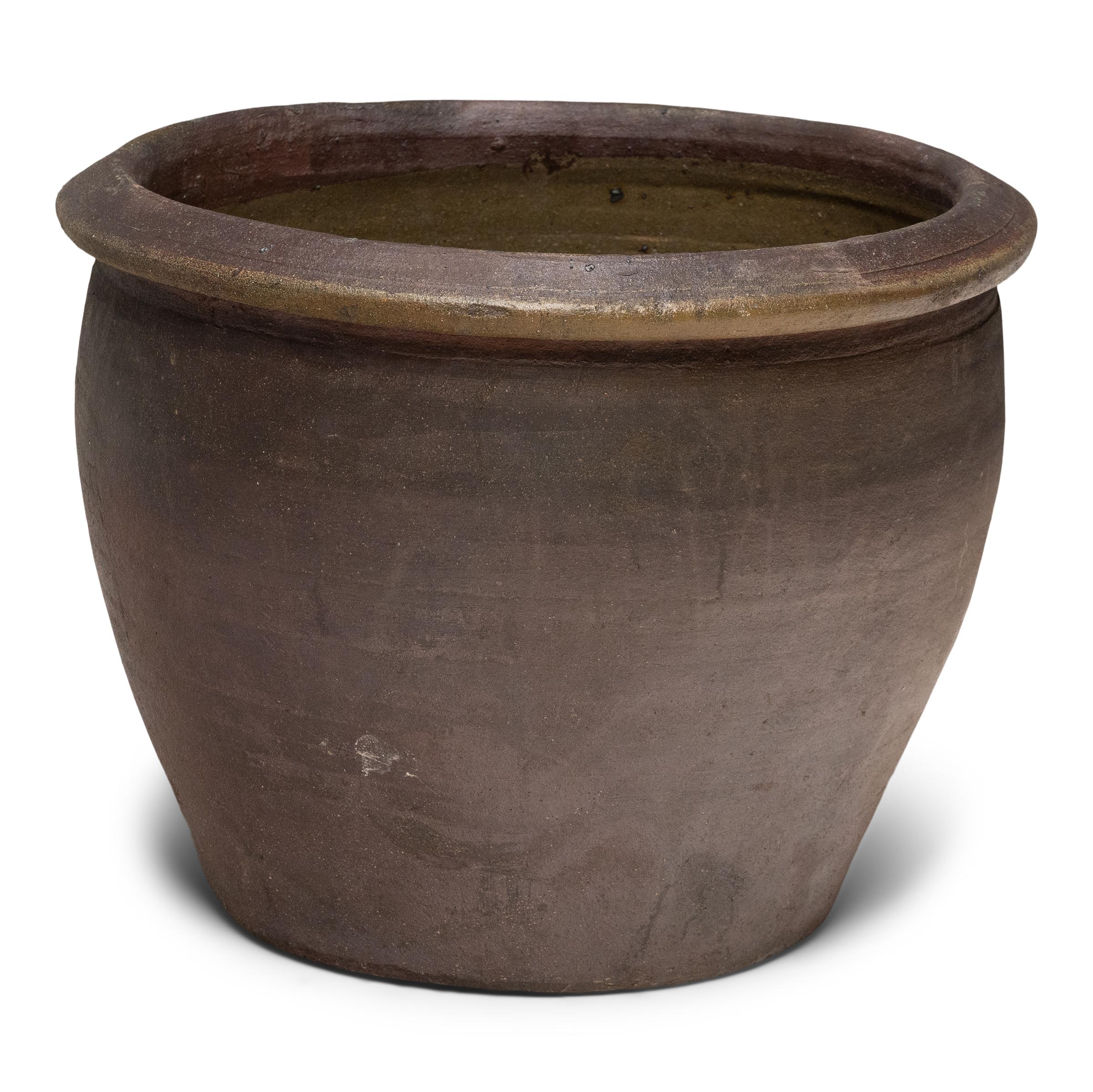Questo laghetto basso in terracotta ha una forma minimale e una finitura splendidamente irregolare. Il vaso ha una silhouette affusolata, con una bocca larga, una base stretta e un bordo semplice e svasato. Lasciato senza decorazioni, l'esterno è