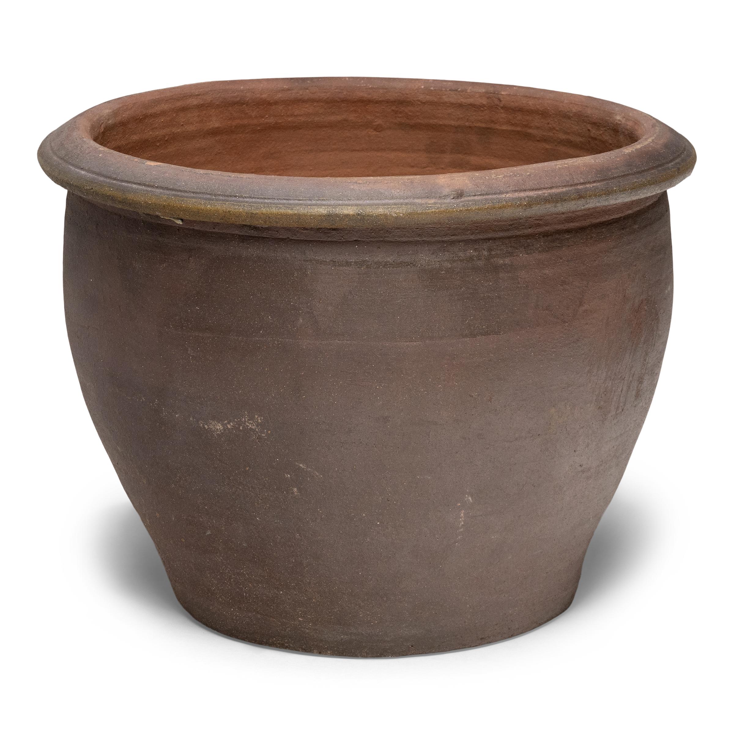 This low terracotta planter sits with a minimal form and a beautifully irregular finish. The vessel has a tapered silhouette, with a wide mouth, narrowed base and simple, flared rim. Left undecorated, the exterior is darkened by a natural ash glaze