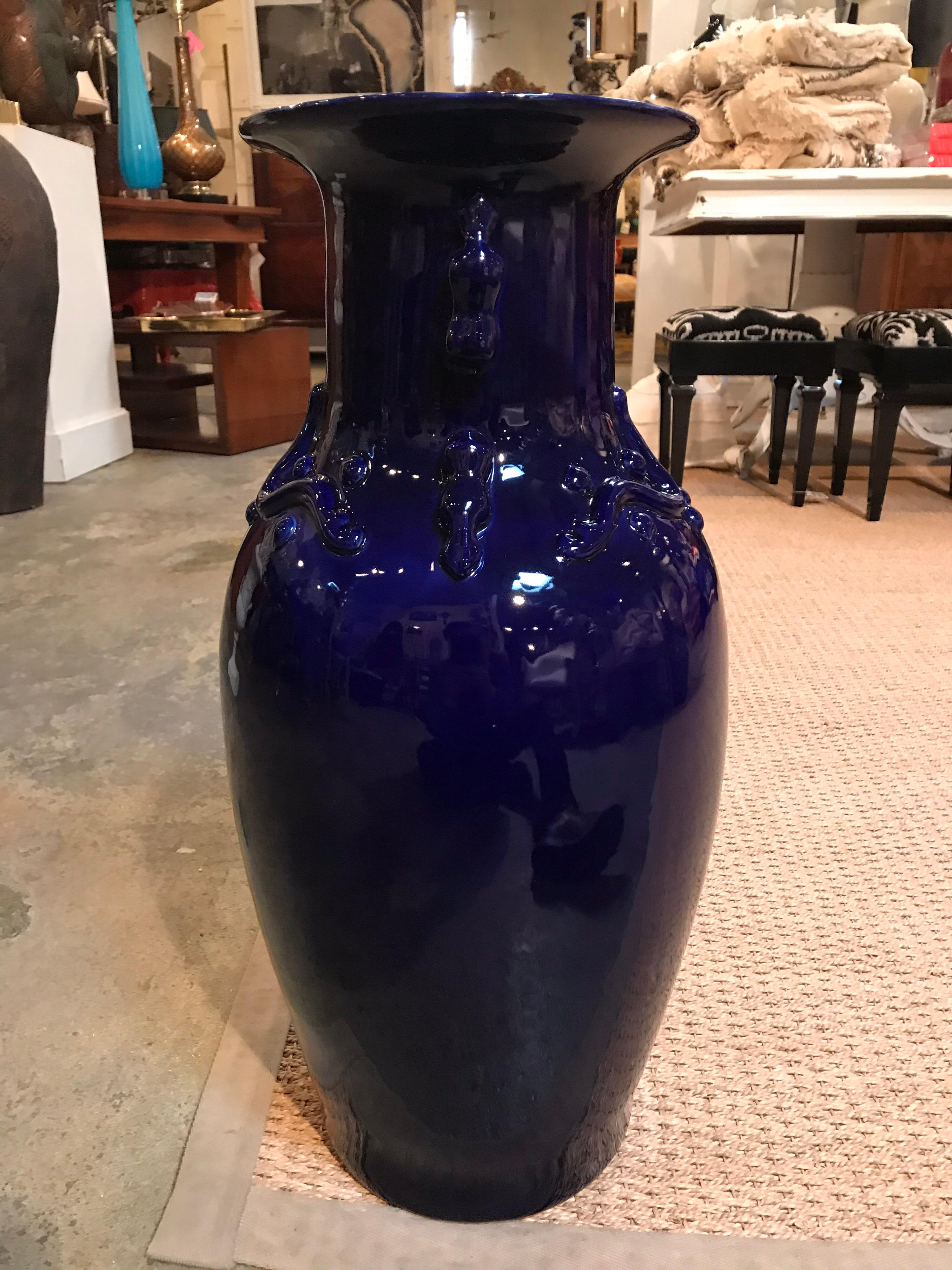 This handmade vase was made in the mid-20th century in a glazed cobalt blue ceramic with decorative raised shapes around the base of the neck. The item is thin and hollow.