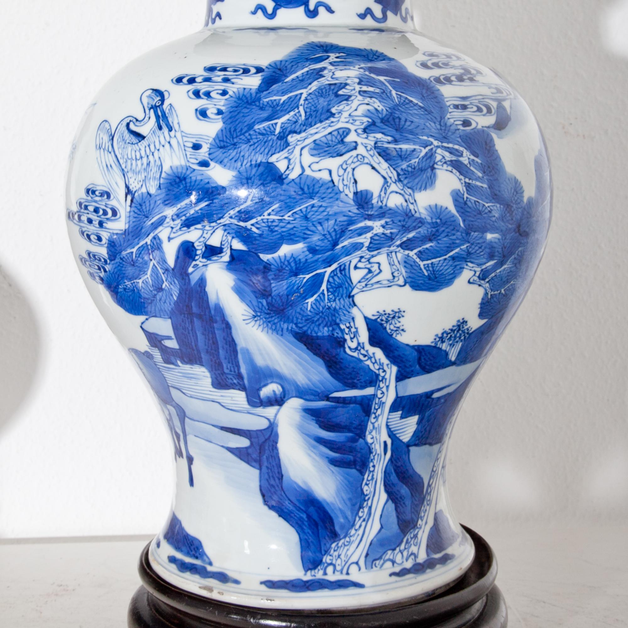 Porcelain vase standing on a round wooden base with openwork wooden lid of the Kang'Hsi period. The bulbous wall with underglaze blue landscape painting with storks and deer. At the upper rim depiction of the treasures, such as the Artemis-leaf and