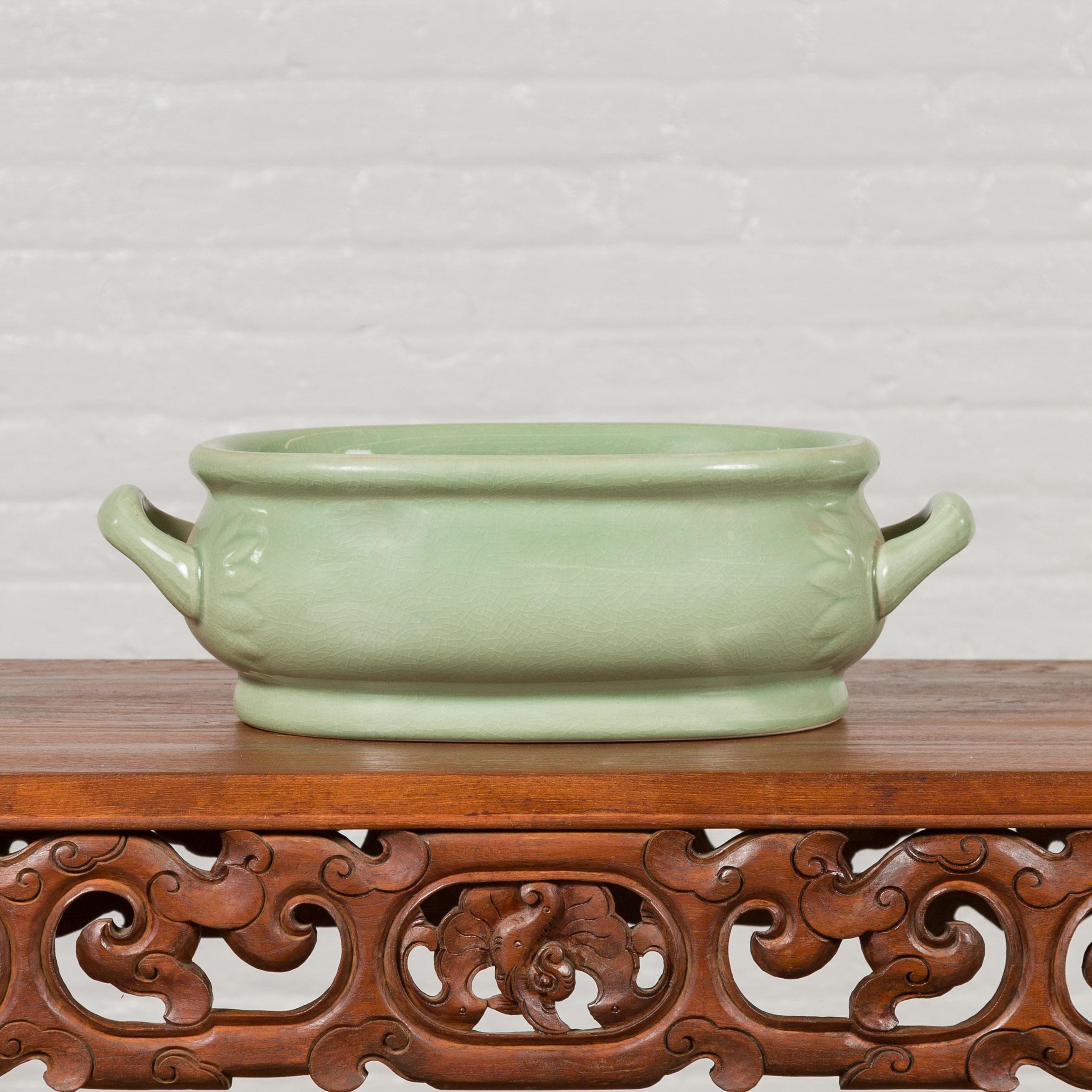 A Chinese vintage celadon foot bath from the mid-20th century, with two handles and foliage motifs. Created in China during the midcentury period, this celadon foot bath features an oval shape topped with a thick lip. The body, showcasing a slightly