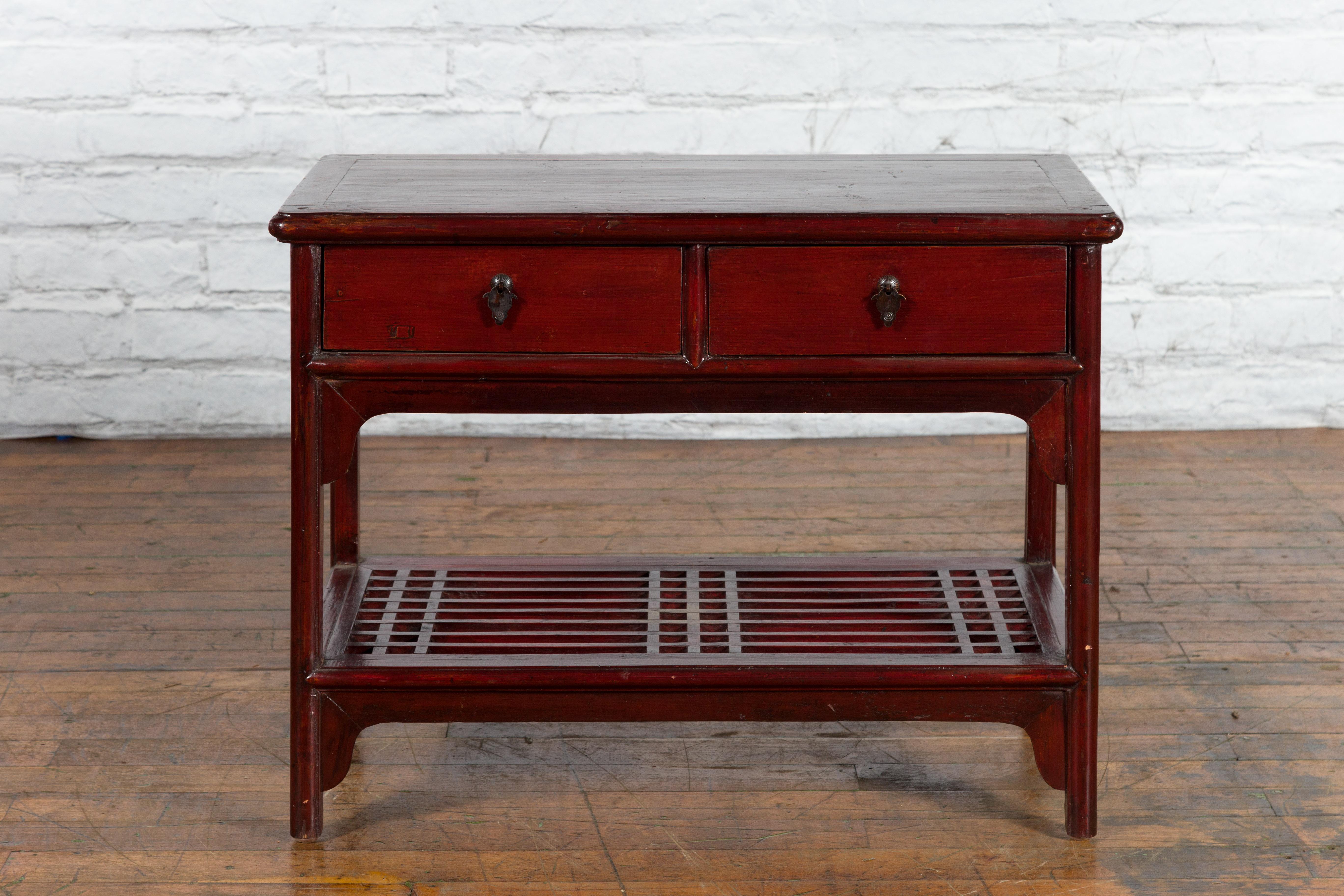 A vintage Chinese oxblood color side table from the mid 20th century with fretwork shelf, two drawers and shaped pulls. Created in China during the Midcentury period, this vintage lacquered side table features a rectangular top with central board