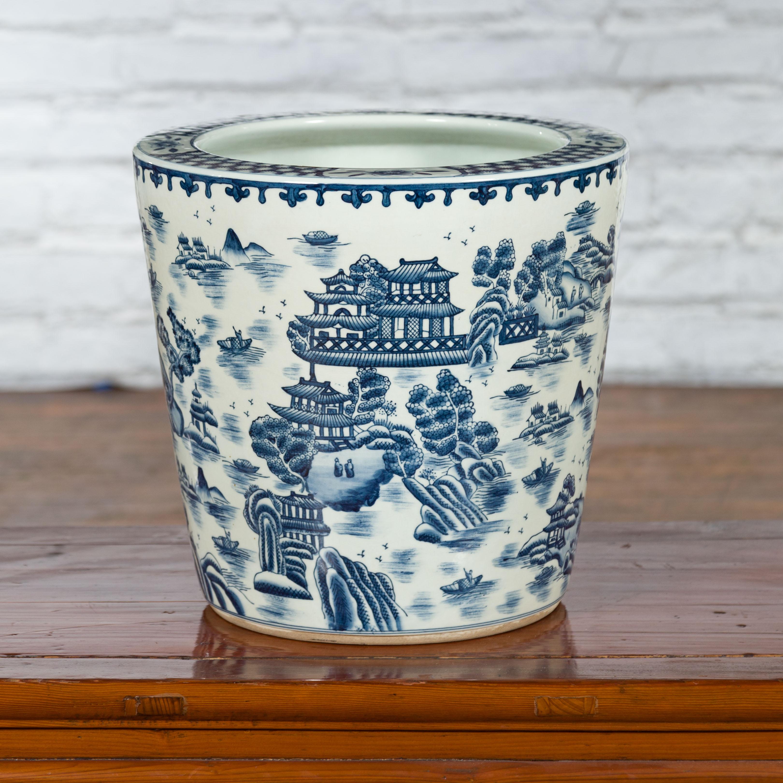A Chinese vintage porcelain cache-pot planter from the mid 20th century with hand-painted blue and white décor depicting landscapes, architectures and figures in boats patterns. Created in China during the midcentury period, this porcelain cache-pot
