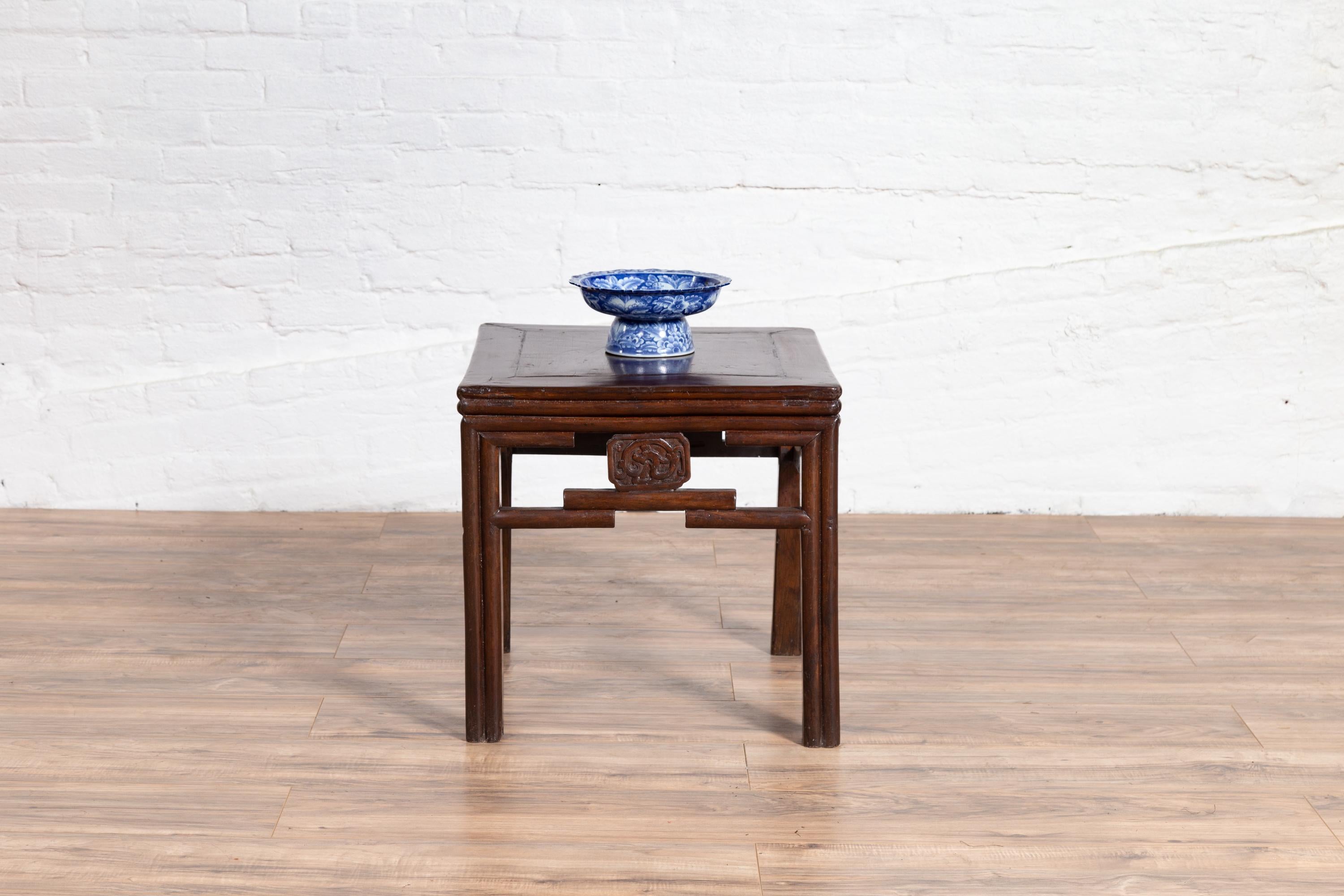 A Chinese vintage Qing dynasty style side table from the mid-20th century, with carved medallions and dark patina. Born in China during the mid-century period, this charming side table features a square planked top sitting above an open fretwork