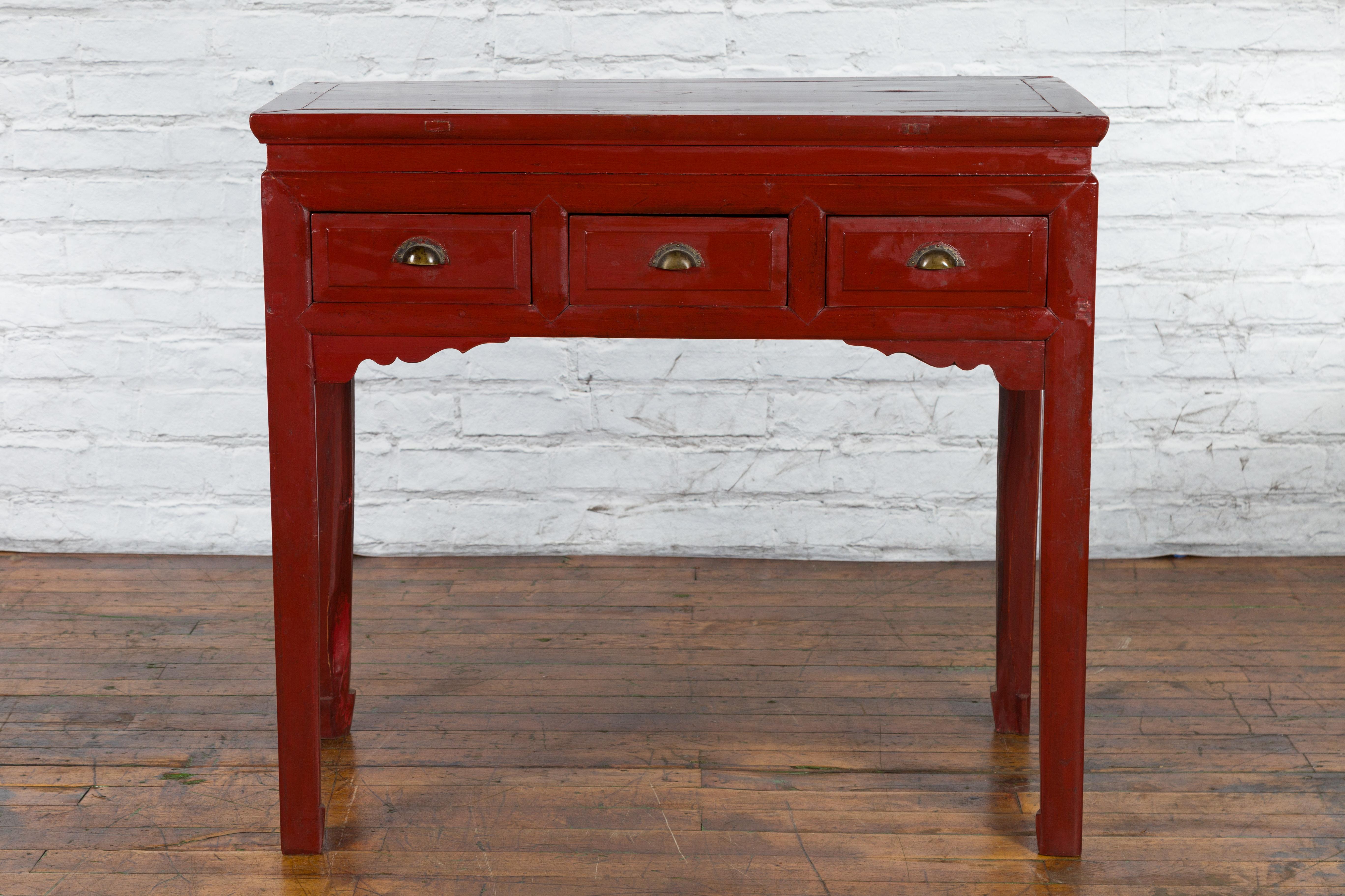 A vintage Chinese red lacquered console table from the mid 20th century, with three drawers, waisted top, carved spandrels and scalloped hardware. Created in China during the Midcentury period, this red lacquered console table features a rectangular