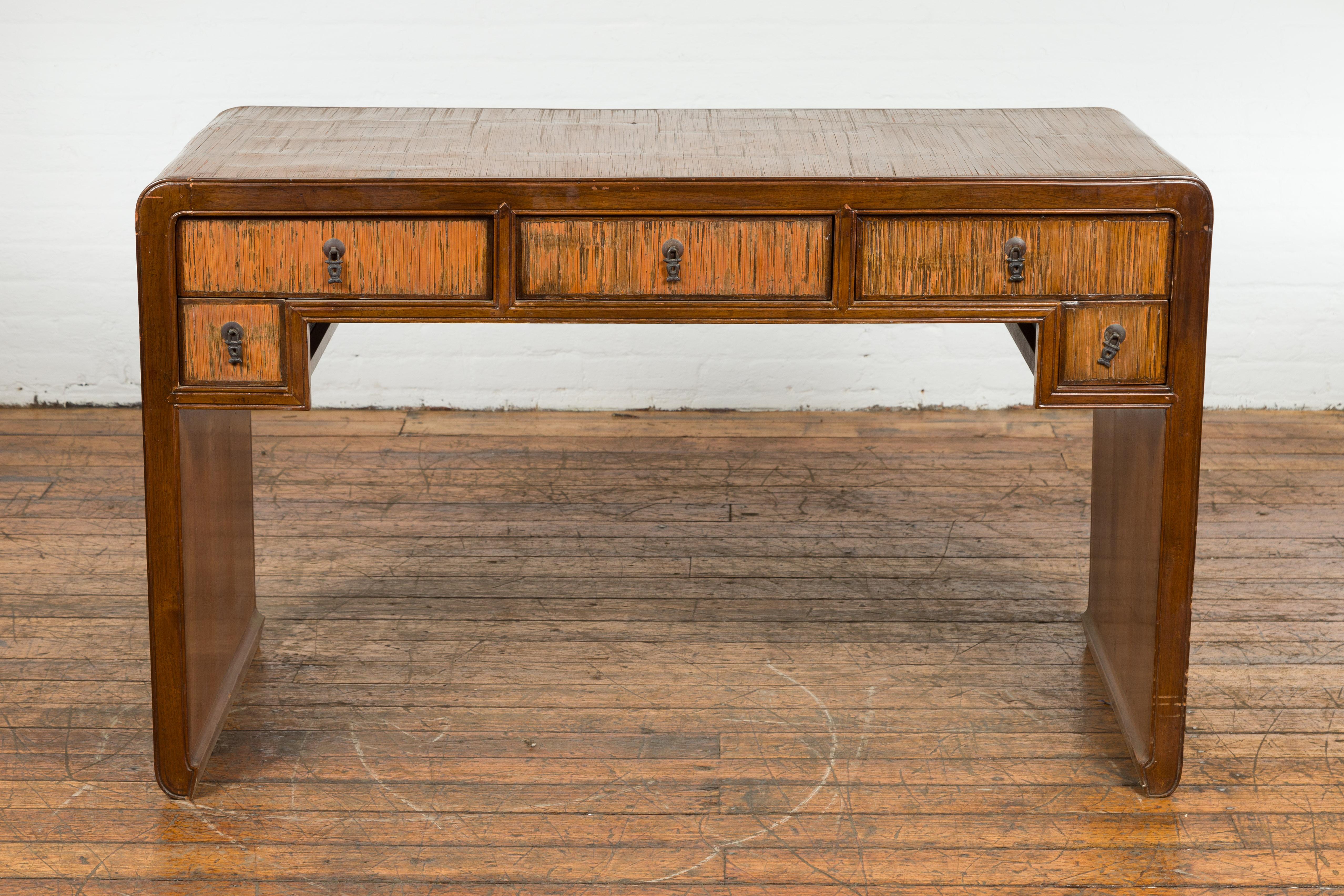 A vintage Chinese waterfall desk with five drawers, opium mat top and rustic character. Emanating a blend of exotic allure and sophisticated craftsmanship, this vintage Chinese opium mat desk is a captivating piece of functional artistry. Born in