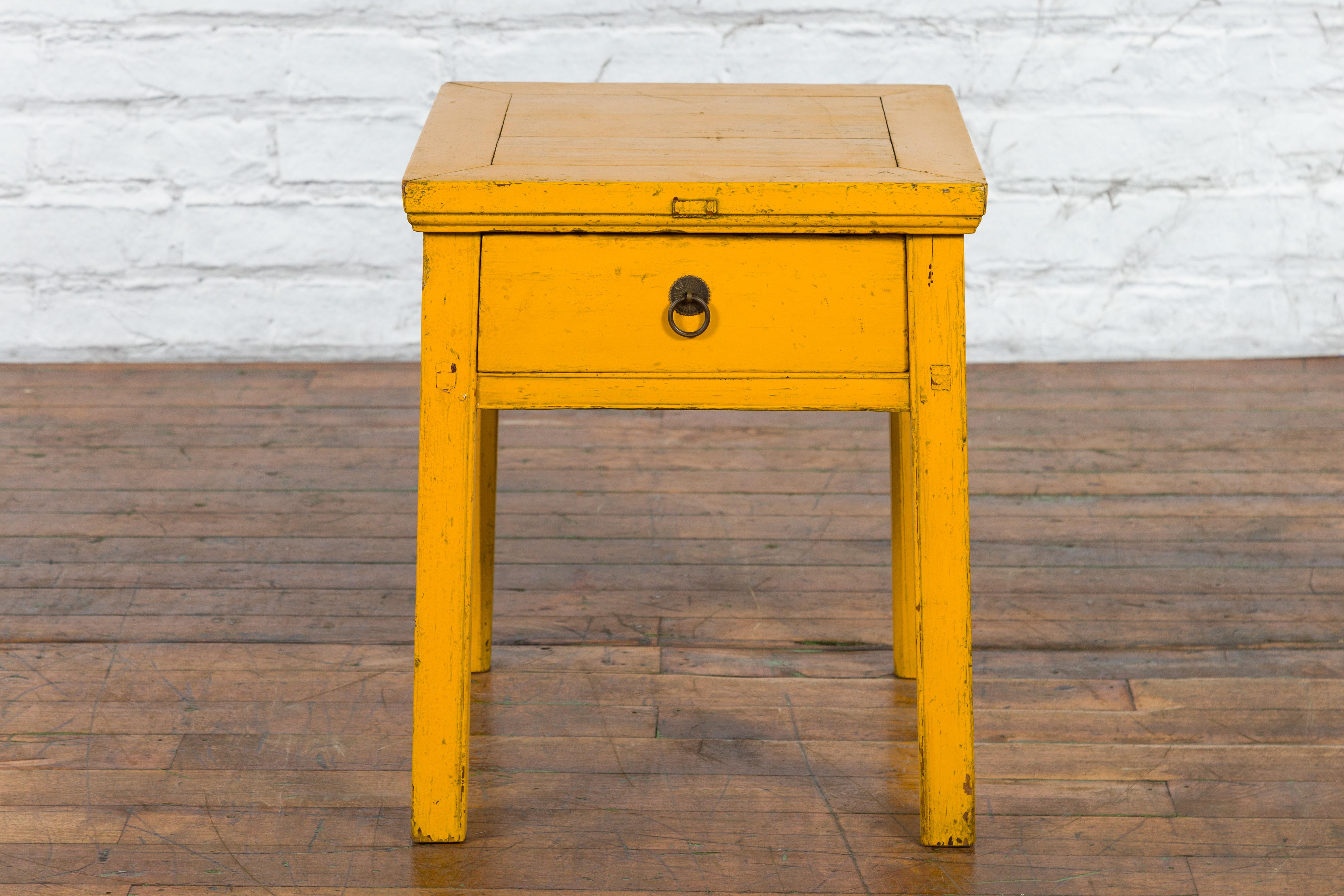 A Chinese vintage side table from the mid 20th century with yellow lacquer, distressed finish and single drawer. Created in China during the Midcentury period, this side table features a square top with central board, sitting above a single drawer