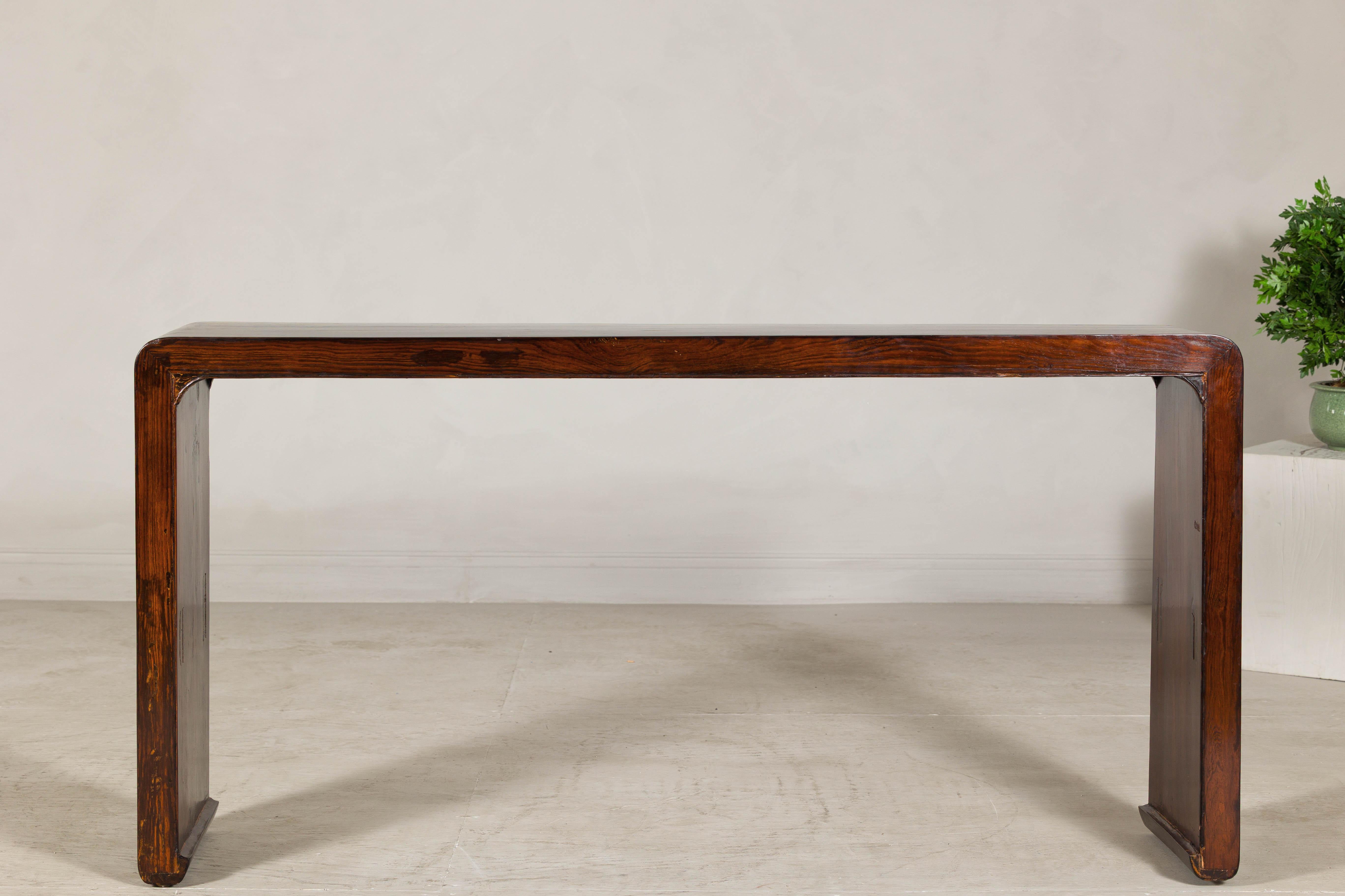 With its fluid silhouette and restrained detailing, this antique Chinese waterfall console table exemplifies the beauty of Ming-inspired minimalism. Crafted in hardwood with a warm, reddish-brown patina, the design is defined by its continuous