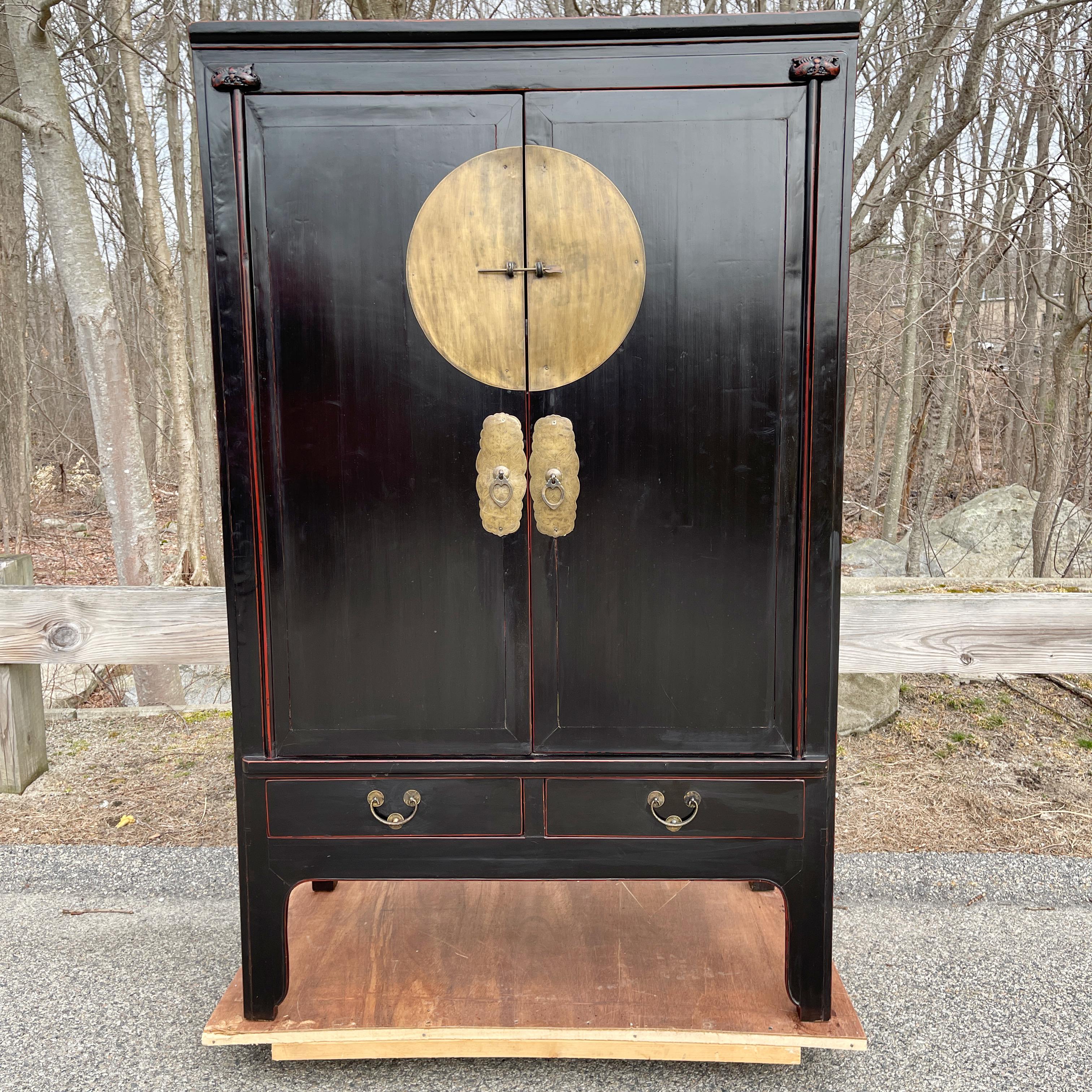 Chinese Qing Dynasty wedding cabinet in black lacquer with red accents featuring large circular brass medallion hardware on two 45 inch high doors above two drawers.
The interior is fitted with a removable shelf above two side by side drawers and