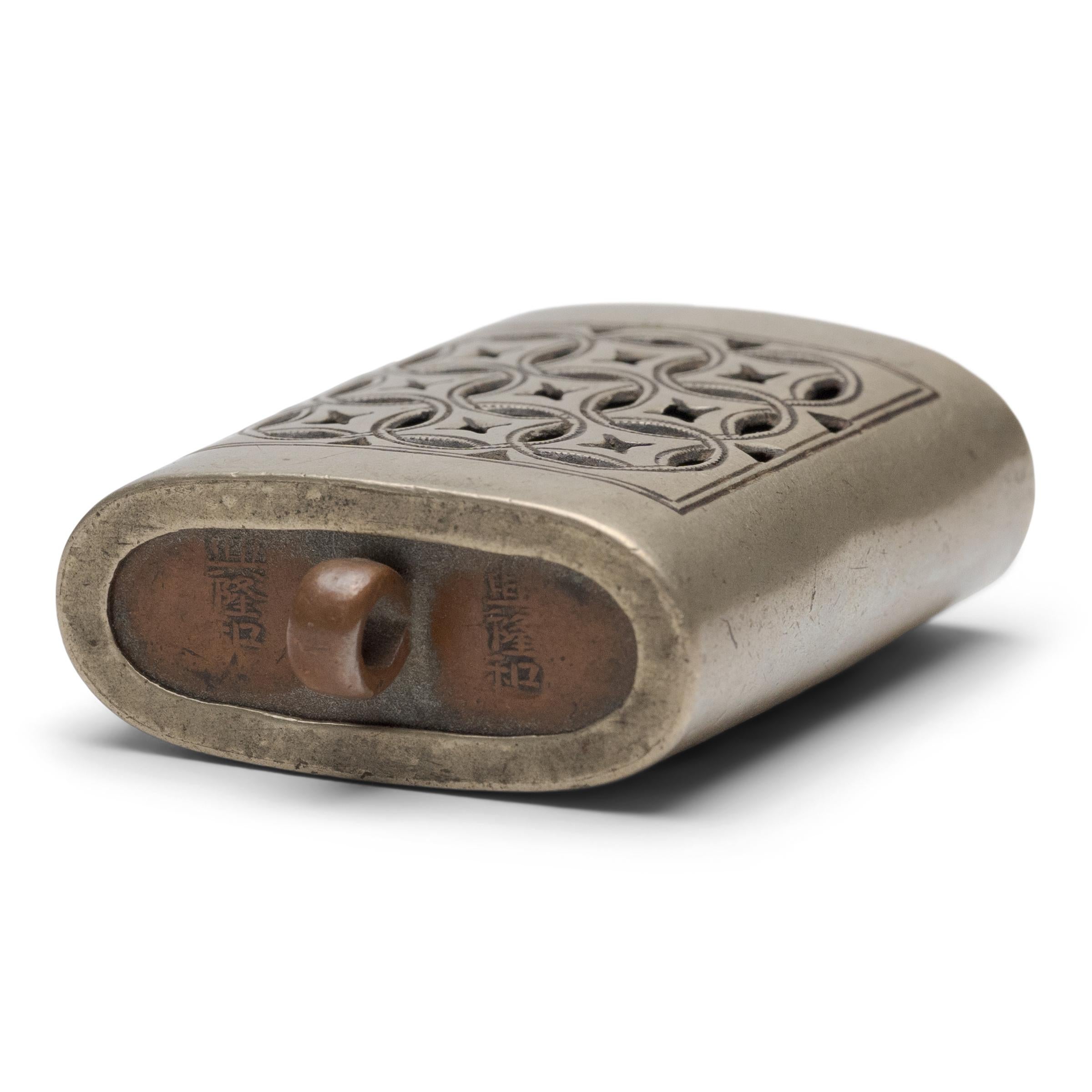 Pequeño toggle de bolsillo de latón blanco y cobre diseñado como caja portátil para la escoria, un recipiente utilizado para recoger la ceniza de opio gastada (escoria). El cierre tiene un diseño perforado de monedas entrelazadas, símbolos de