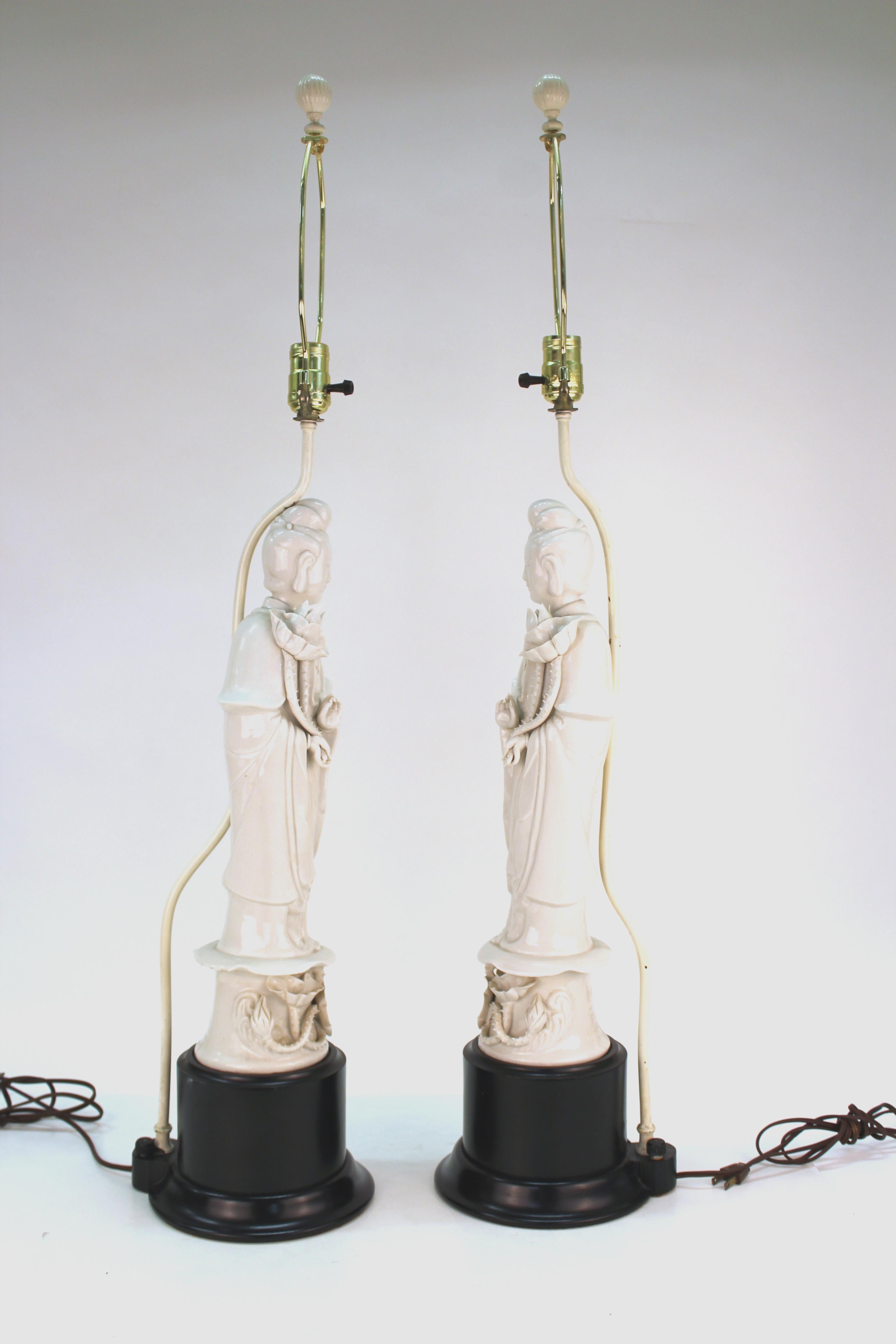 Pair of Chinese Export Guanyin Buddha table lamps in white ceramic atop black circular bases. The pair has Hong Kong marks on the back. There is some chipping to the finish on the metal pipes behind the Buddhas holding up the bulb sockets. In