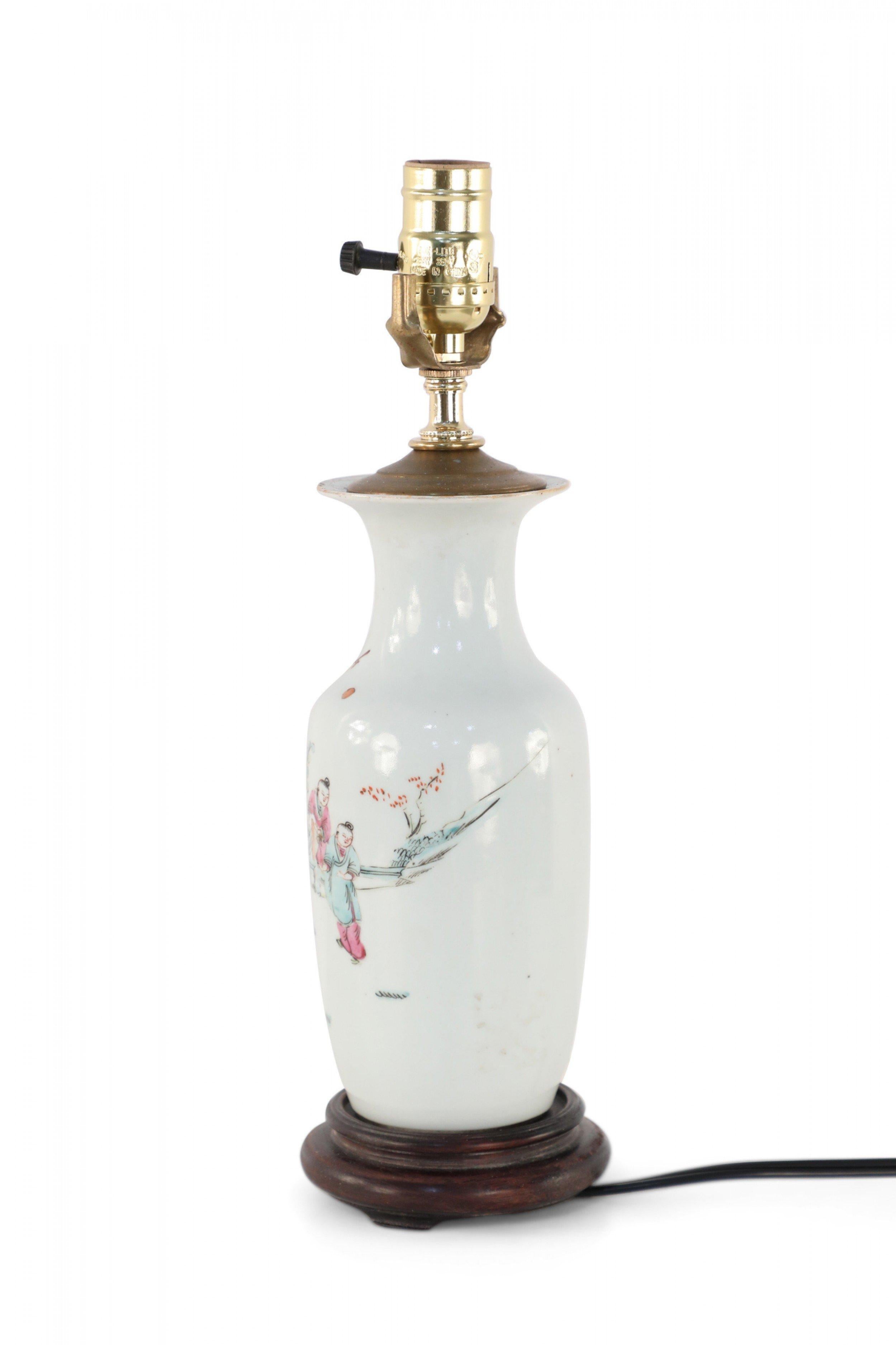 Antique Chinese (Early 20th Century) table lamp made from a white porcelain urn painted with a scene of children playing in orange, pink, and red, mounted on a brown wooden base and fitted with brass hardware.
 
