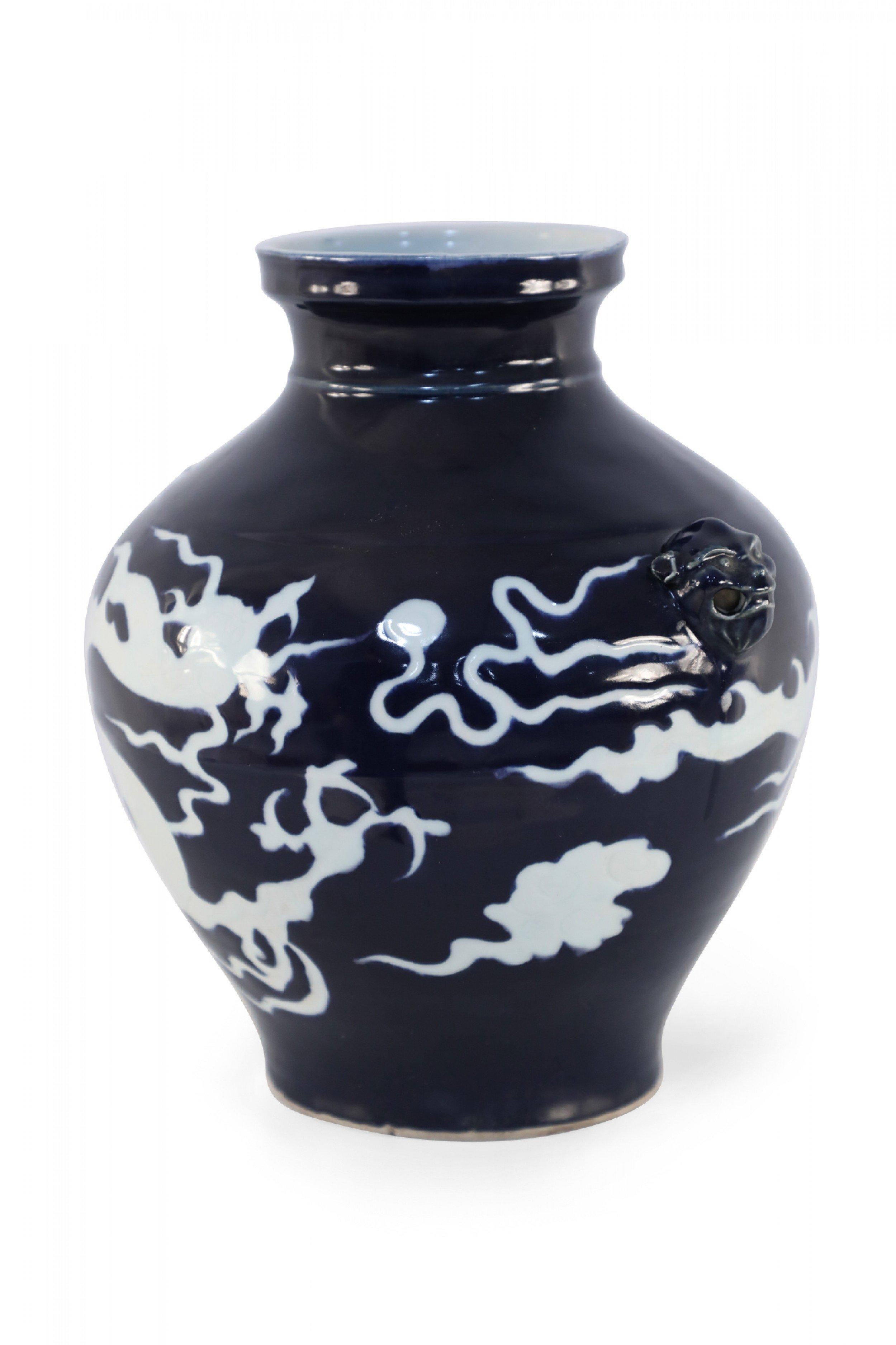 Chinese bulbous, dark blue porcelain pot crafted in the Yuan Dynasty style, wrapped in the dynamic white shape of a dragon and accented with foo dog ornamentation along the sides.
  