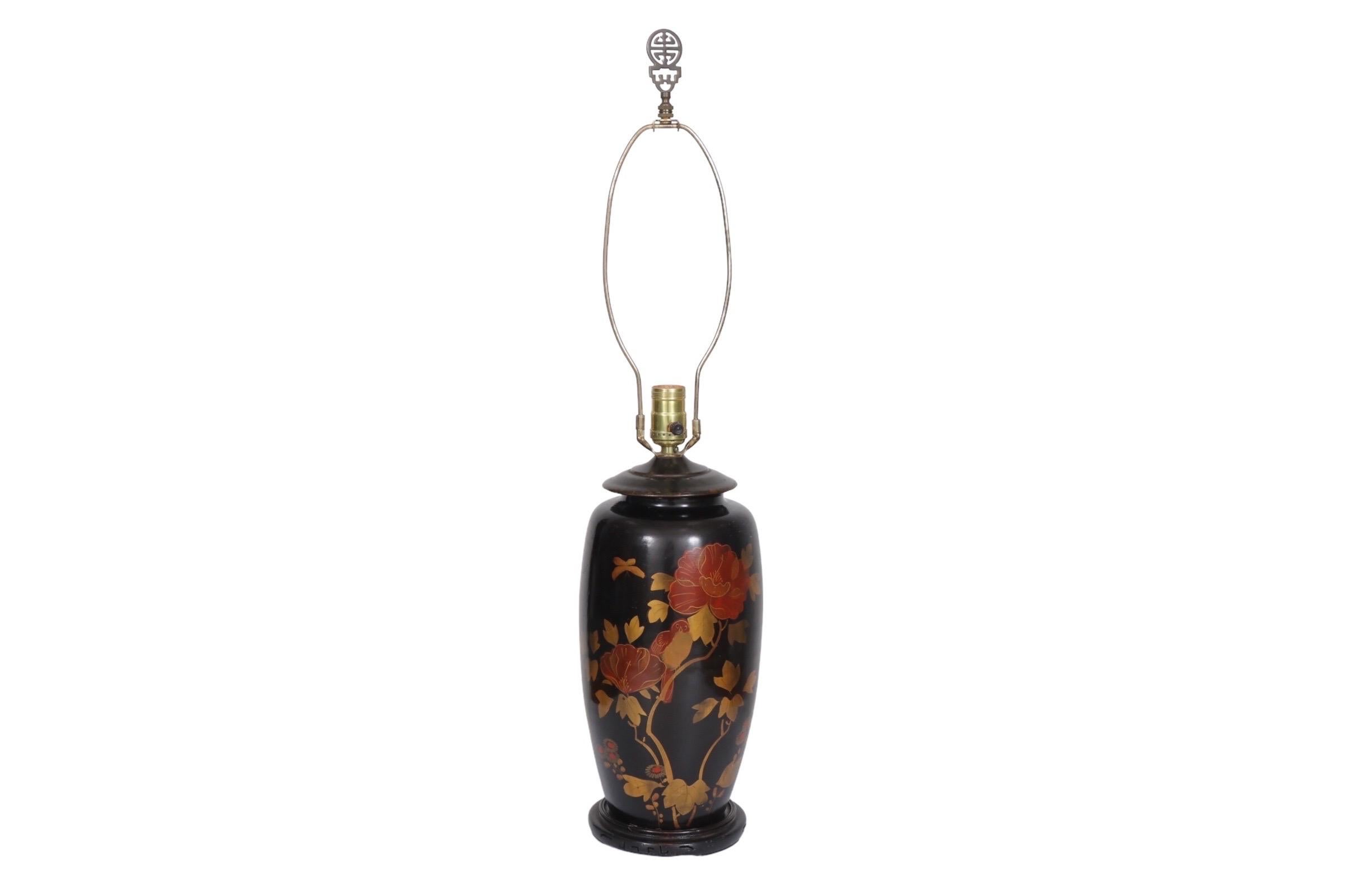 A black Japanned Chinoiserie table lamp. The urn shaped vase has a metal cap and is exquisitely decorated on the front with hand painted red camellia flowers, gold foliage and birds. Sits on a round stepped wooden base. The brass finial is the