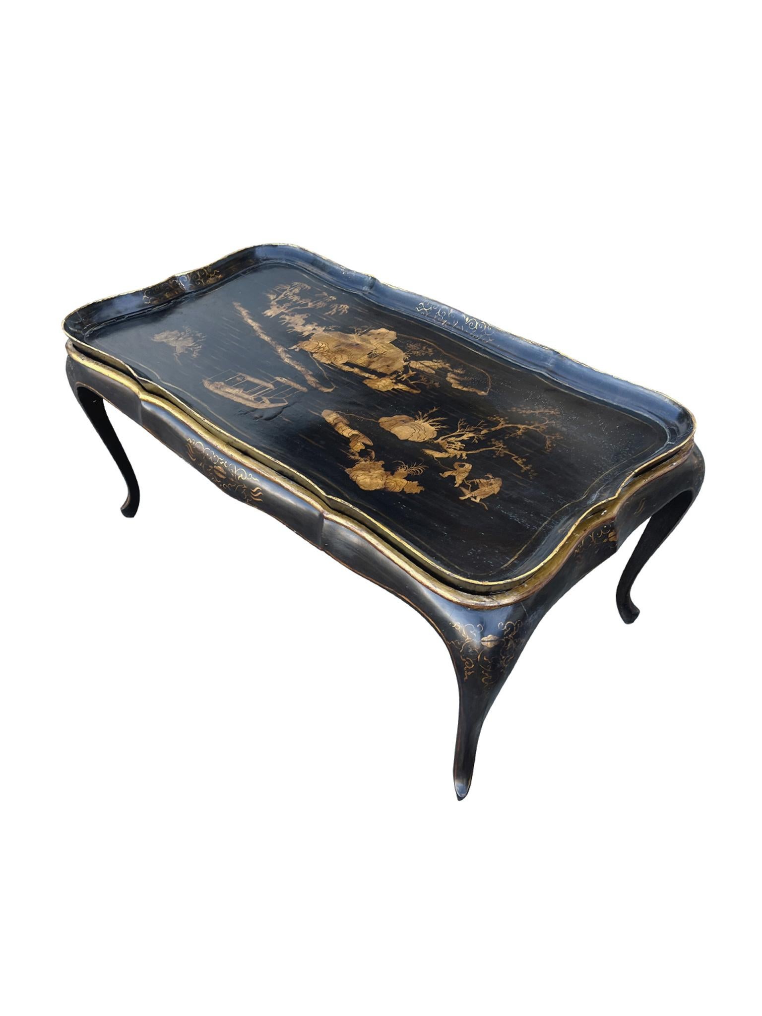Realizzato negli anni '30, questo tavolino a vassoio in legno a cineserie è composto da un piano e da una base a vassoio staccabili, entrambi in una ricca lacca nera e con decorazioni dorate a pacchi. Un paesaggio dorato dipinto a mano adorna la