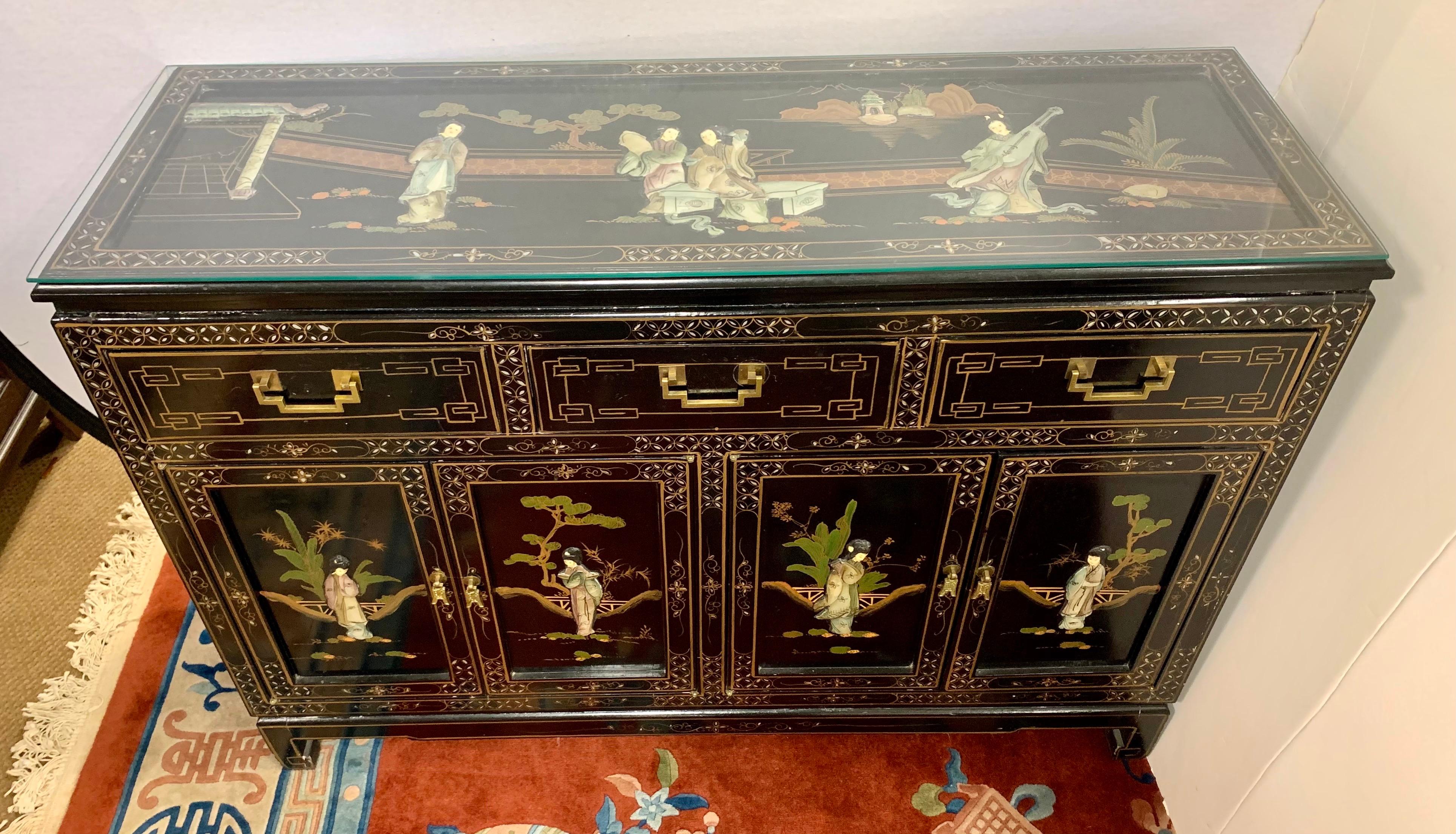 Vintage Chinese black lacquered credenza features carved hardstone figures in a landscape on the tops and doors. Handpainted gold details all around. Sides have a handpainted bamboo and leaf motif. Has three drawers over four doors that open to