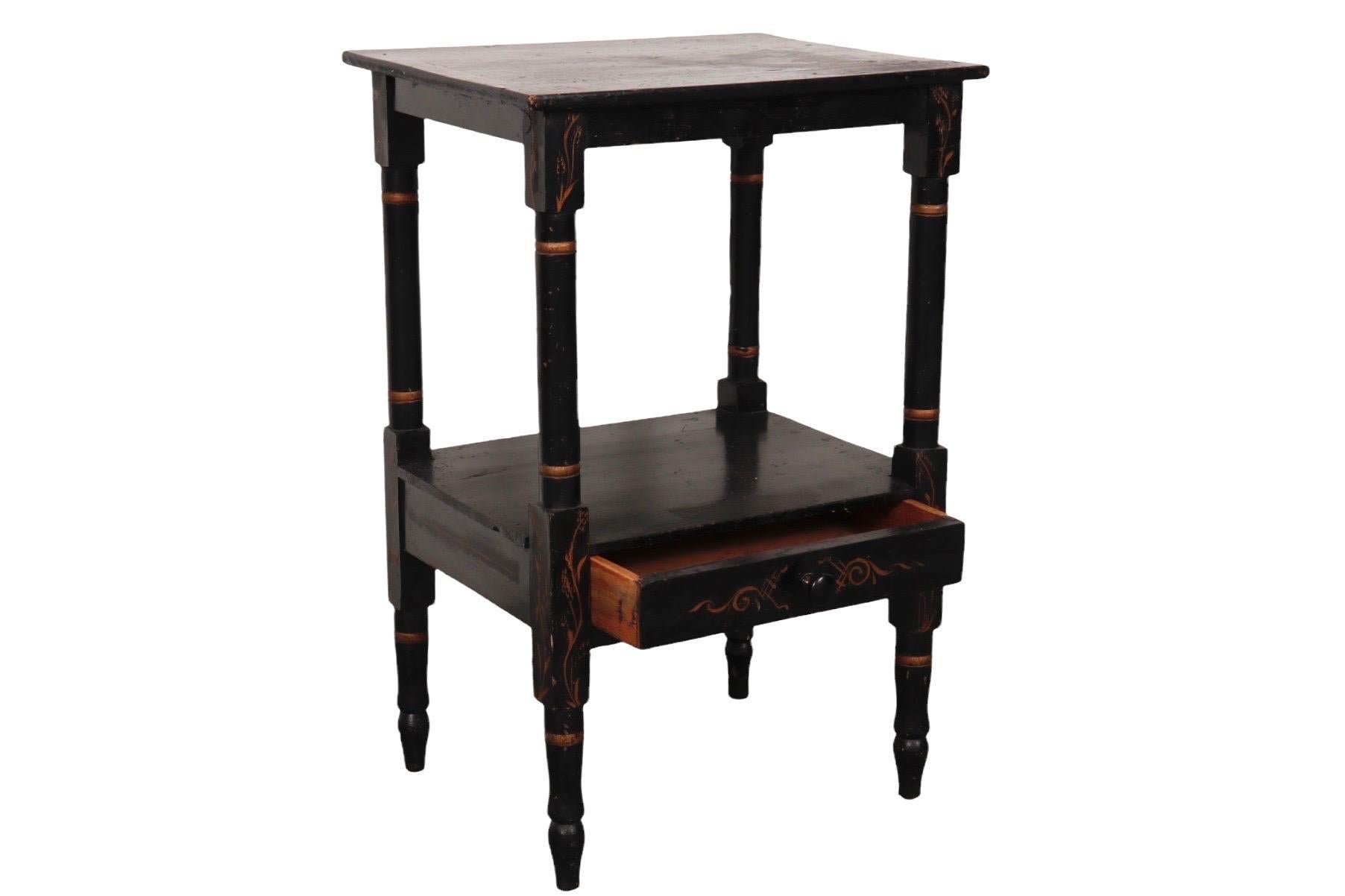 A Chinoiserie side table with a single drawer. Hand painted black and decorated with a simple plant motif and scrolled details in gold. Turned legs terminate in blunt arrow feet.