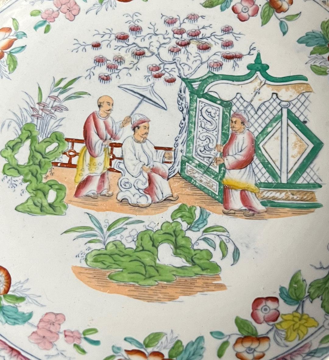Des accents verts vibrants sur cette assiette représentant une scène de jardin semblent montrer un empereur en train de se faire ombrager. Fleurs jaunes et roses pâles sur l'ensemble de la pièce. Quelques traces d'or sur le bord. Date de 1868 au