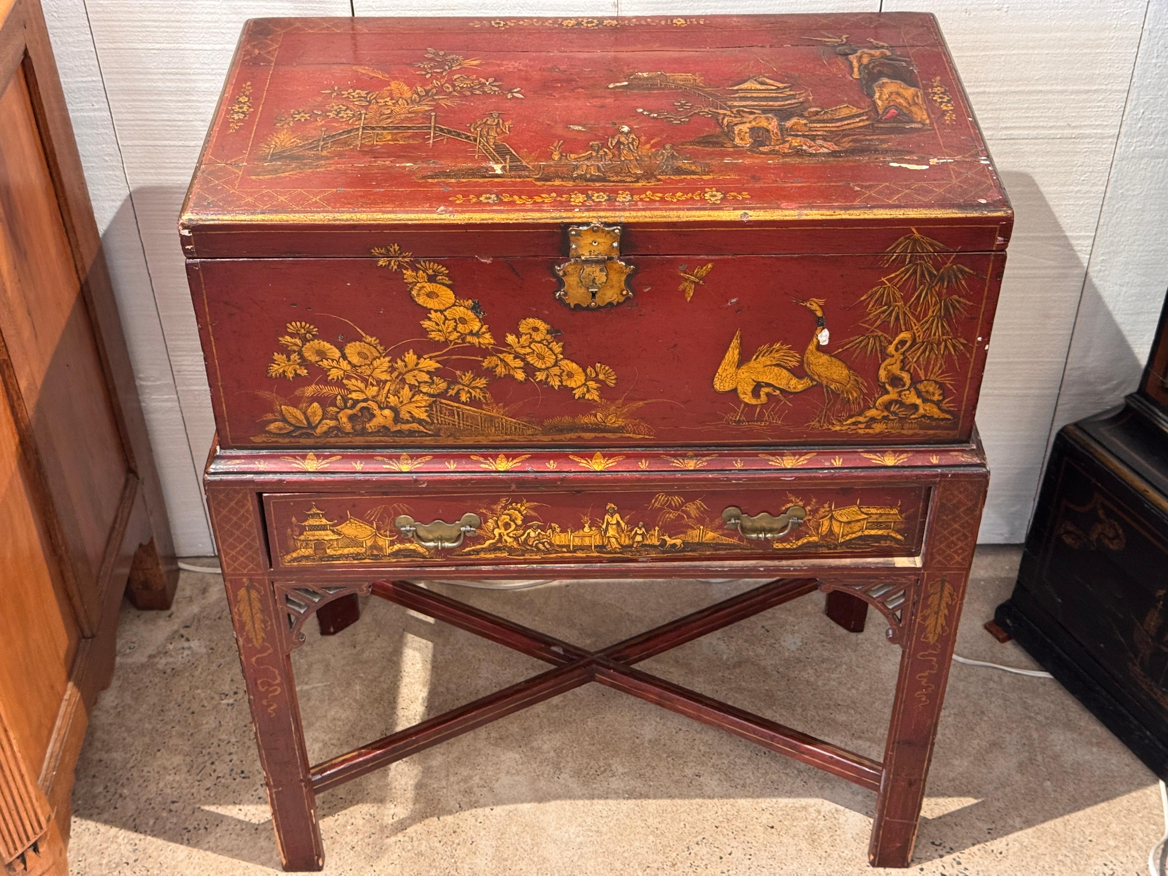 A lacquered box on stand decorated in the chinoiserie taste, featuring gilt landscape and figural scenes on a deep red ground. The box rests on a fitted base with a single drawer and pierced corner brackets joined by an X-form stretcher. The surface
