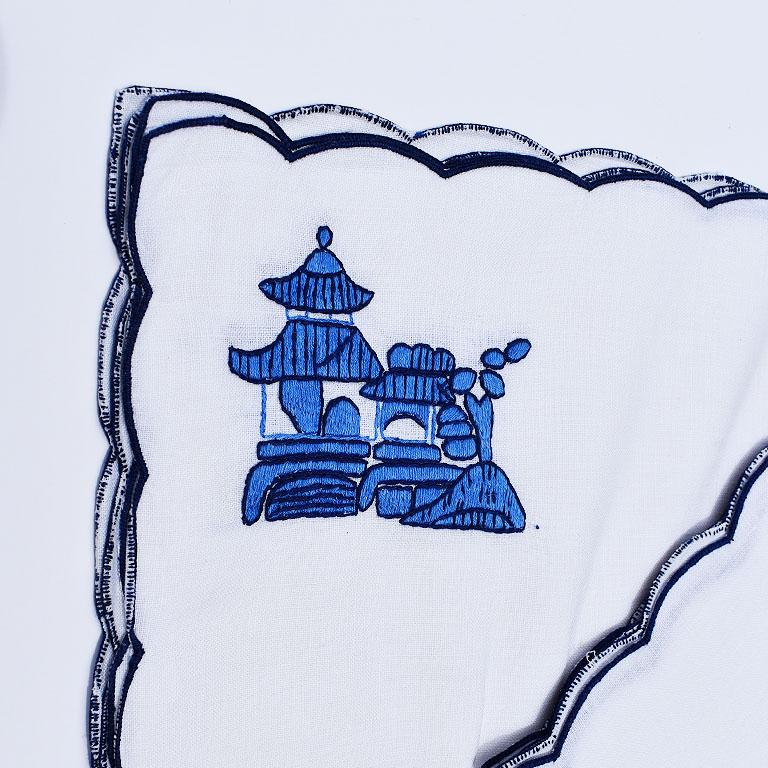 A beautiful set of four chinoiserie blue and white embroidered placemats and matching napkins. The edges of both the cloth napkins and cloth placemats are scalloped with blue embroidery atop a crisp white linen. The placemats feature a pagoda scene