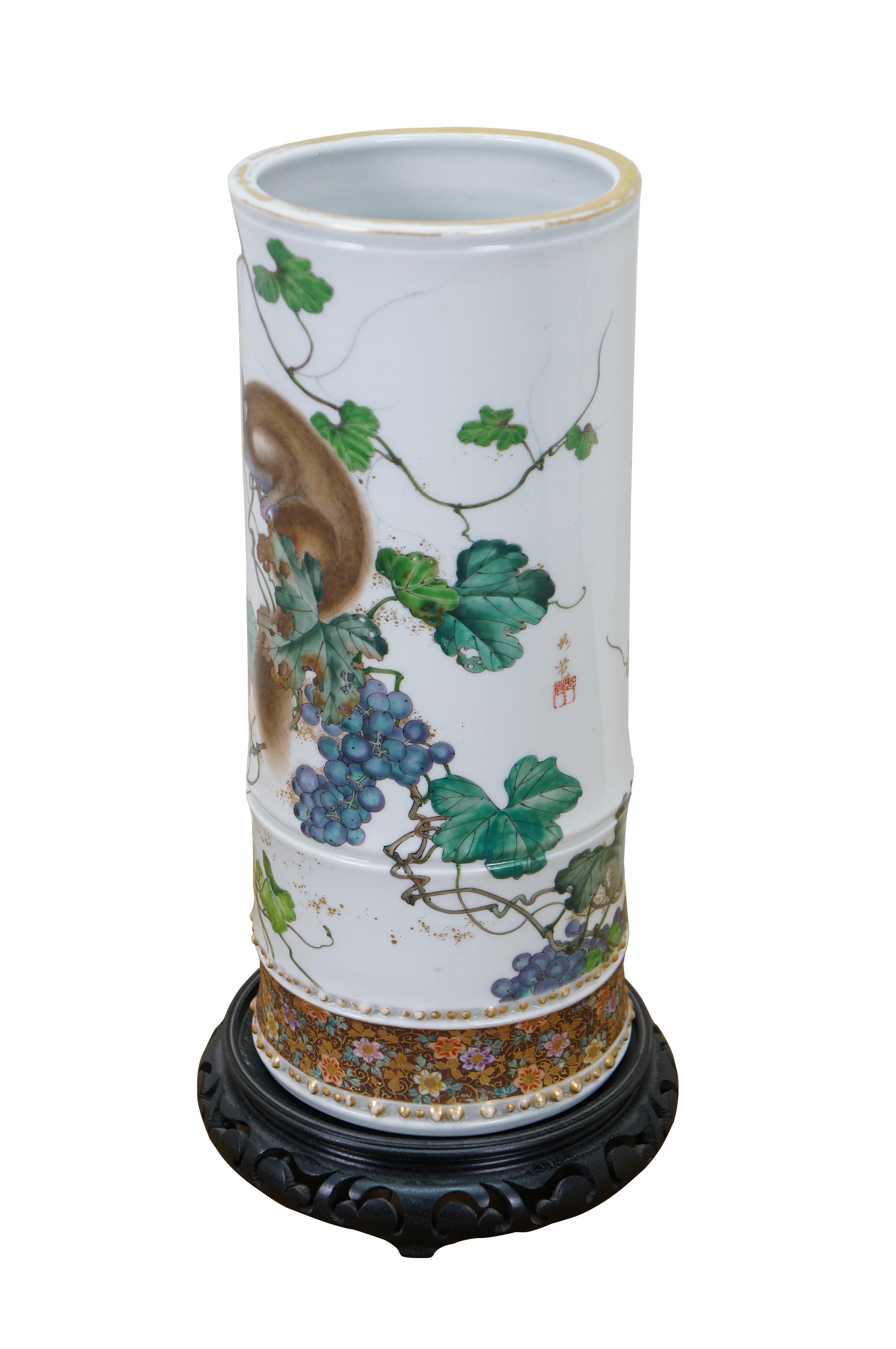Vintage Chinese famille rose porcelain cylindrical vase, decorated with a squirrel perched on a fruiting grape vine, with a dense floral band around the base. Gilt accents. Signed on one side and marked on base. Includes wooden