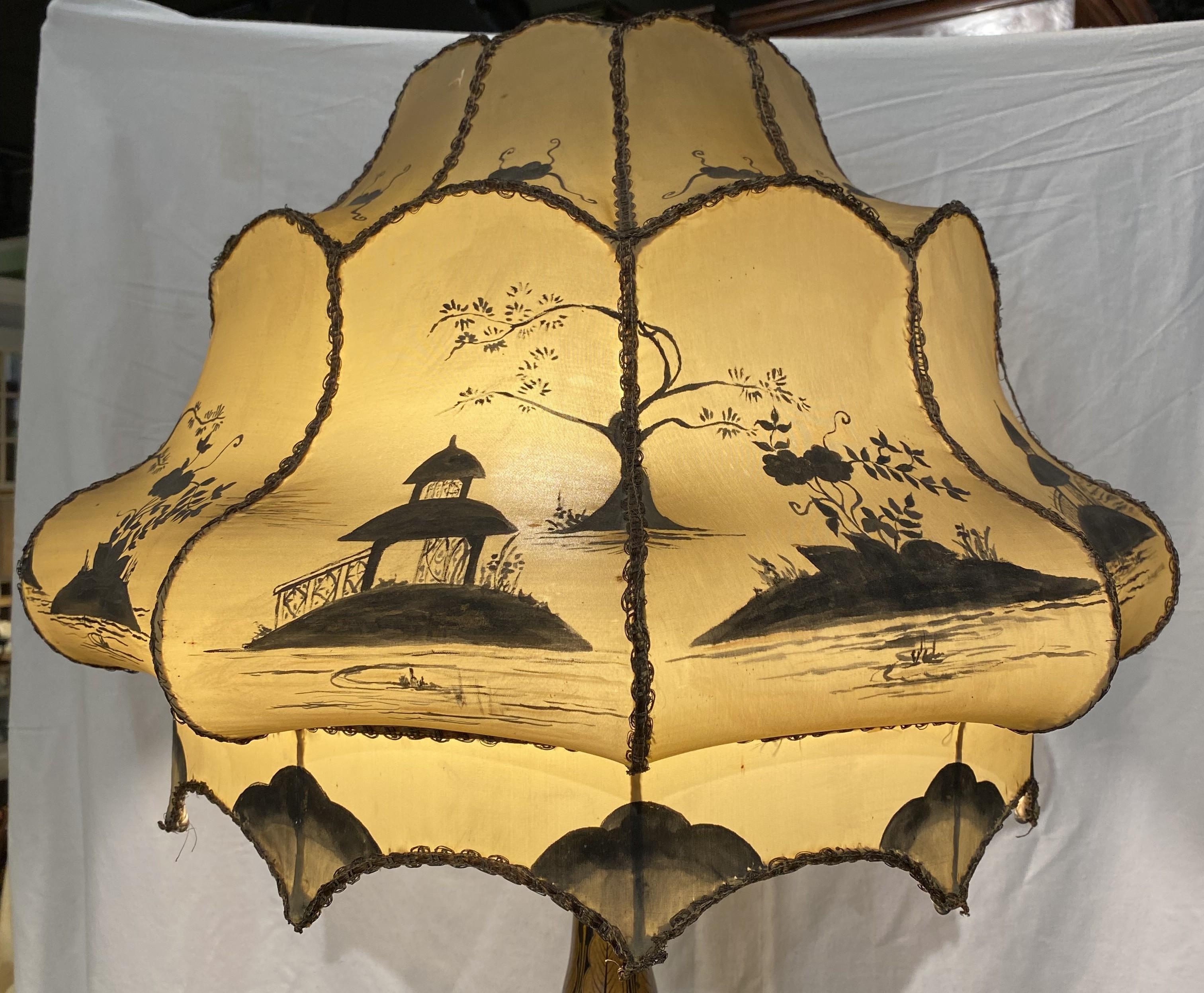 A wonderful Chinoiserie floor lamp with shaped linen hand painted shade and polychromed lacquered round table base featuring relief carved landscape and folkloric scenes with figures, buildings, birds, and fauna. The stem of the lamp continues to