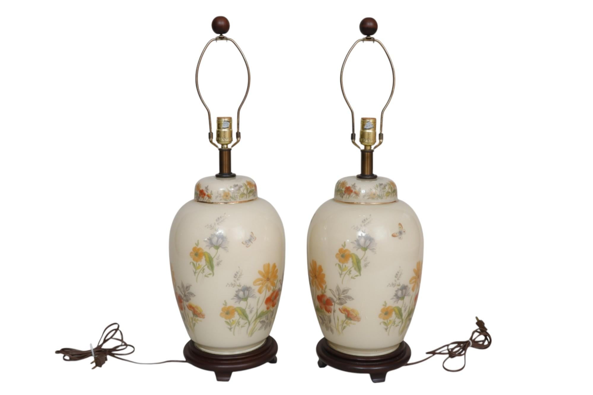 A pair of Chinoiserie glass table lamps. Wooden ball finials secure empire shaped concertina folded shades. Ginger jar shaped vases are painted cream on the interior, with delicate transfers of wildflowers and butterflies in primary colors. Finished