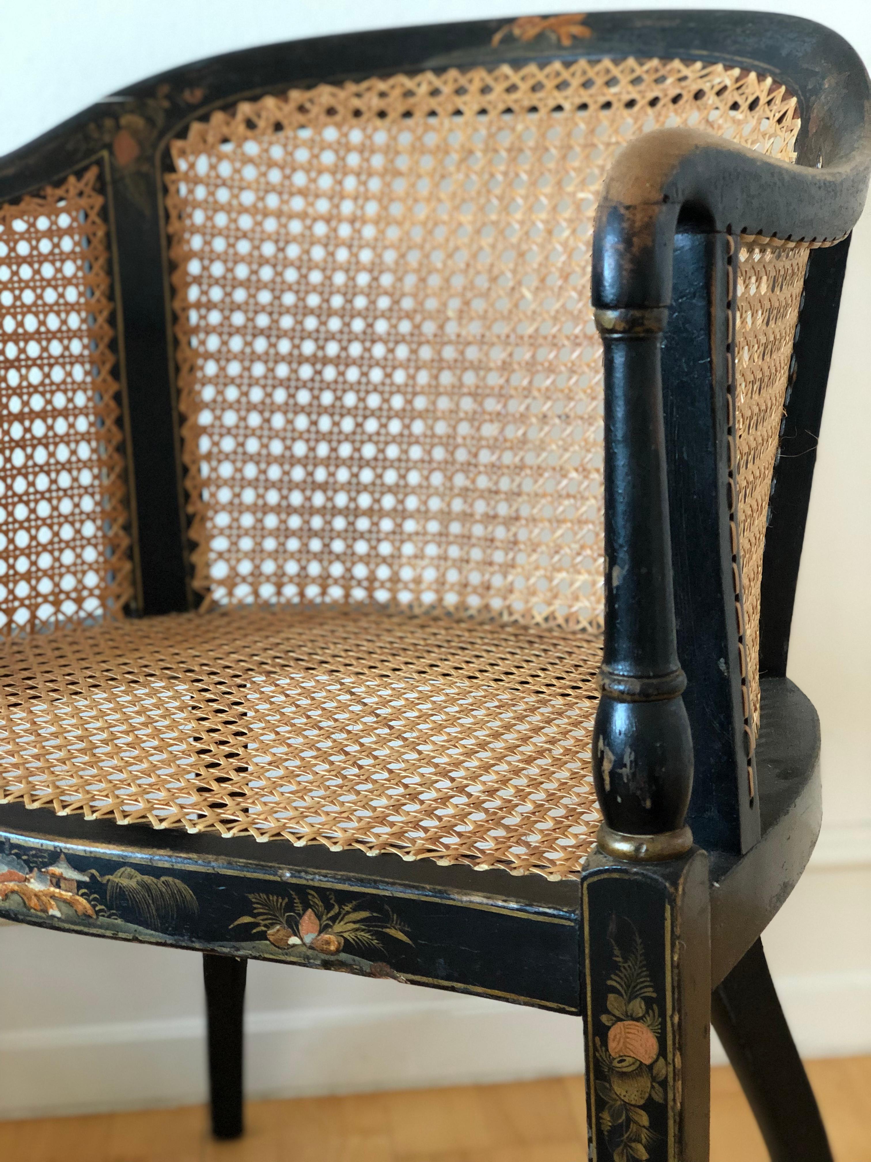 This early 20th-century Chinoiserie tub chair, crafted in the United Kingdom, features a cane seat and back for both comfort and style. The chair is made from black lacquered wood, decorated with gilt Chinoiserie motifs inspired by Chinese and East