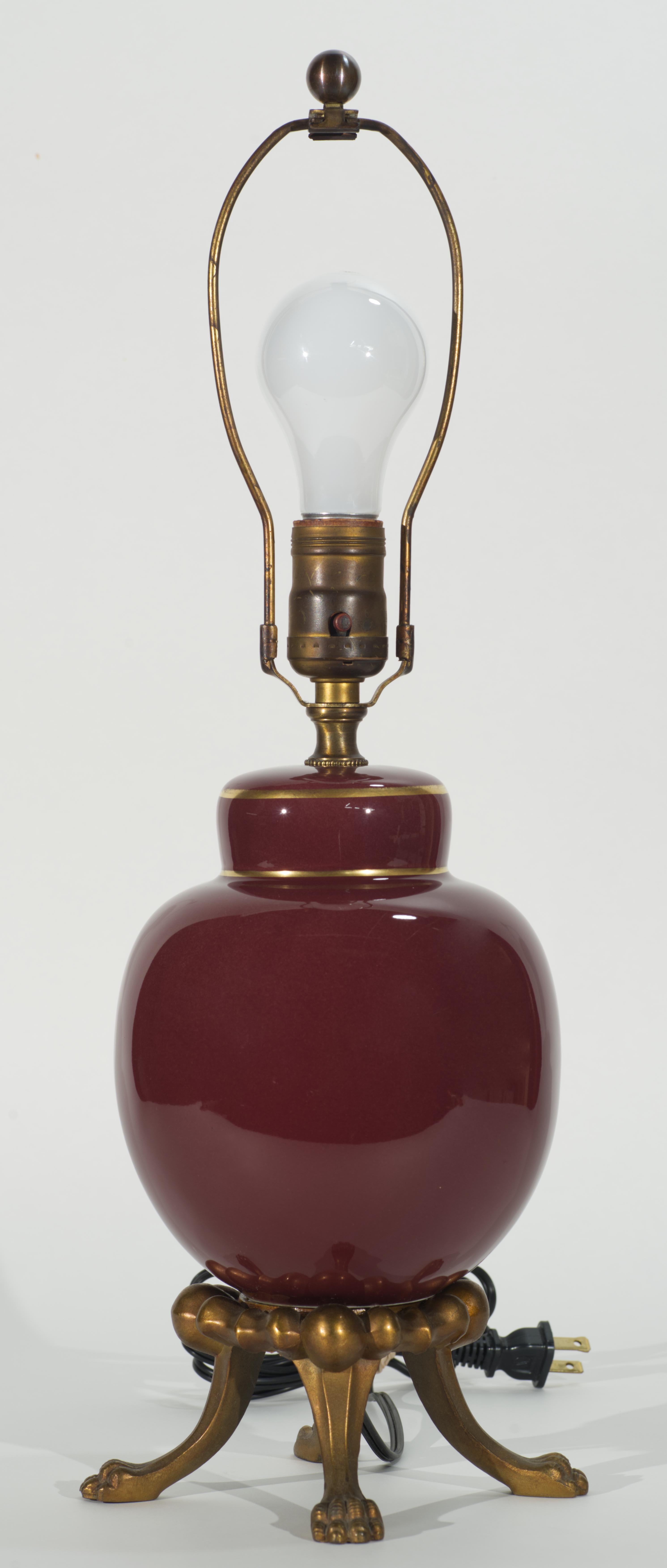  Vintage or antique accent or table lamp features the classic ginger jar or urn shaped body mounted on footed pedestal. The ornate bronze pedestal is standing on 4 highly detailed claw feet. The ceramic body of the lamp is decorated with oxblood
