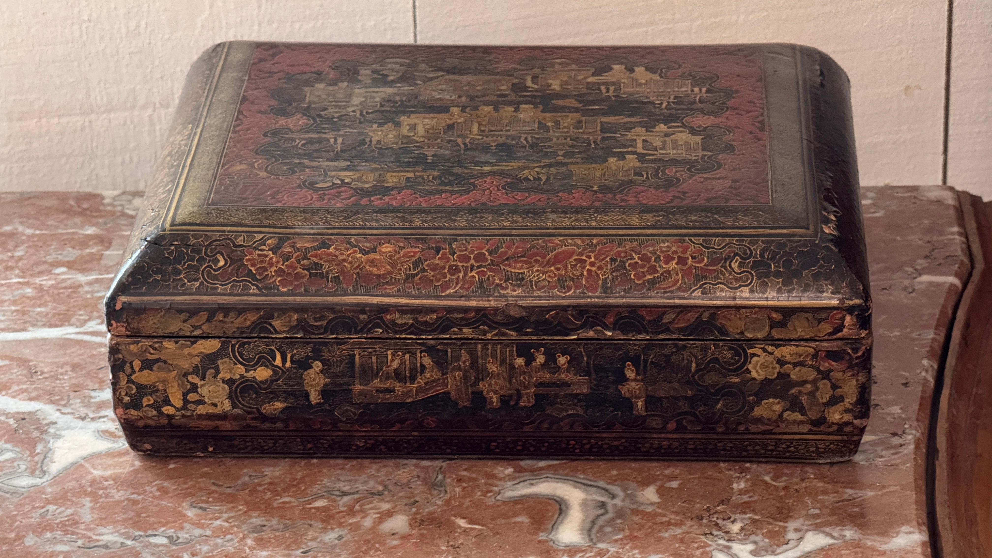 Hand-painted lacquer box with red and gilt Chinoiserie decoration across the lid and sides, showing landscape scenes, figures, and floral borders. The interior retains its original glossy black lacquer. Wear to the edges is visible in the detailed