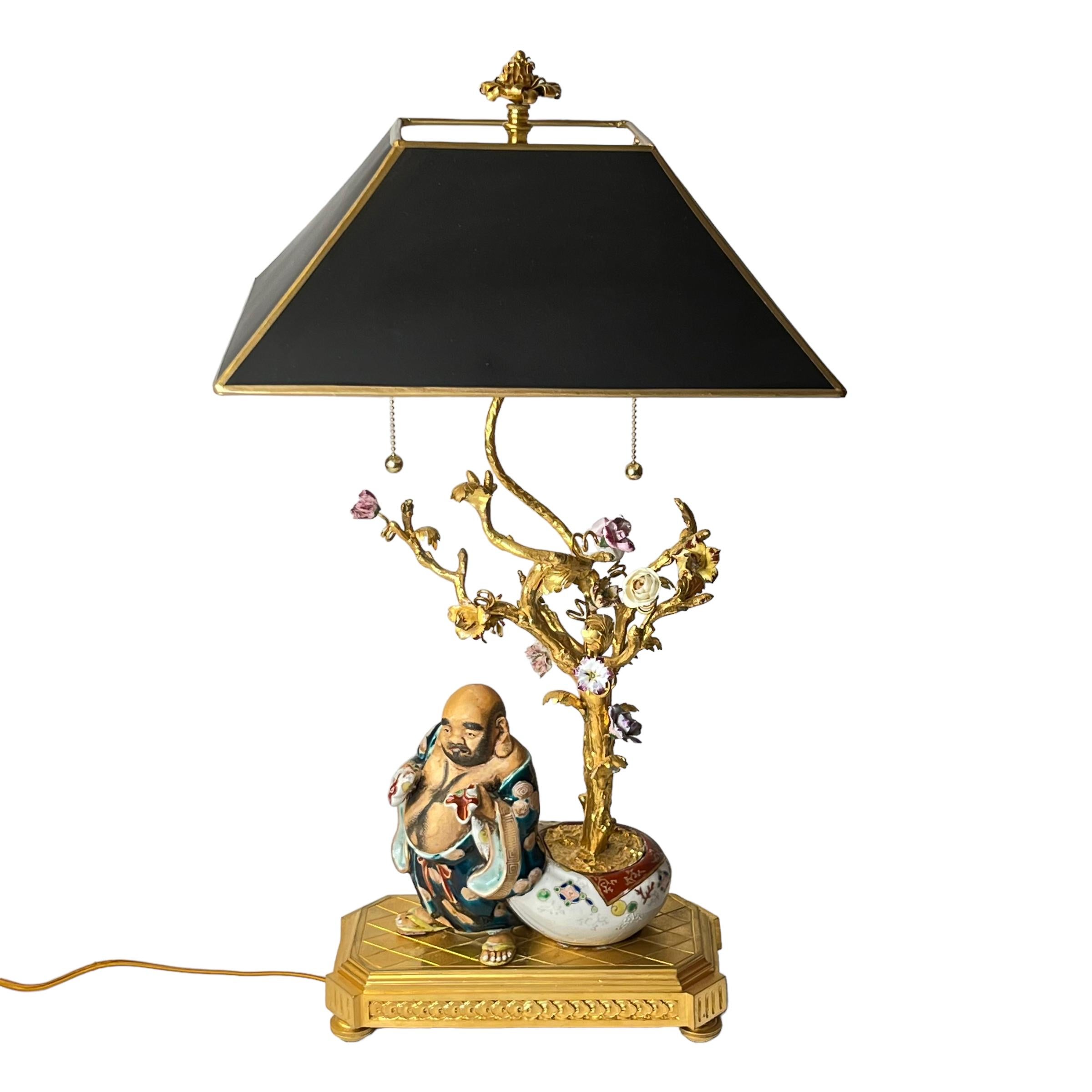 Our lovely table lamp features the polychrome decorated porcelain figure of the laughing Buddha standing beside a tree with porcelain flowers planted in porcelain jardiniere, and a finely cast and gilded bronze stand. Believed to be of French