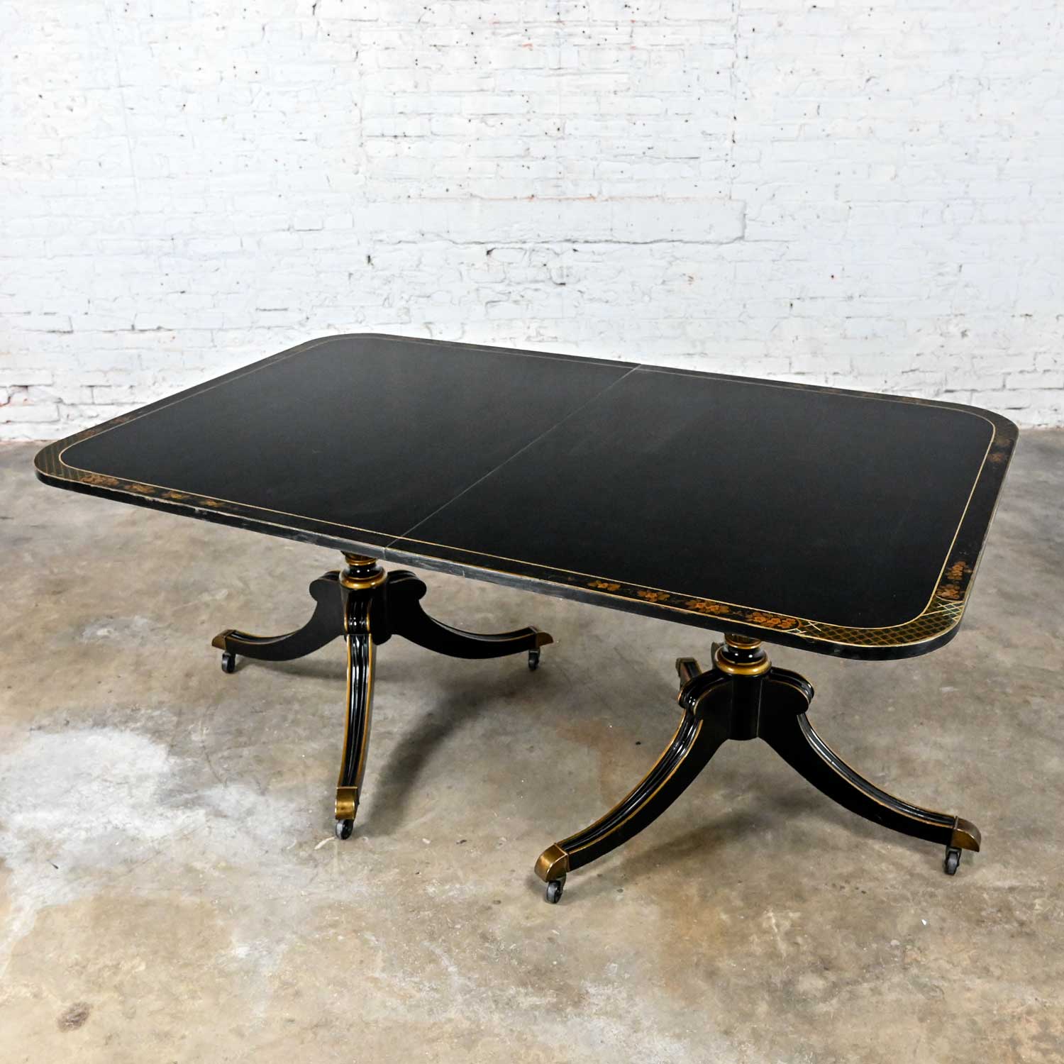 Fantastic vintage Chinoiserie Regency style Union National Inc. dining table with black painted & gilt edged top, 3 skirted apron leaves, & pineapple double pillar tripod base with casters. Painted design is signed on tabletop by J. Sheldon.