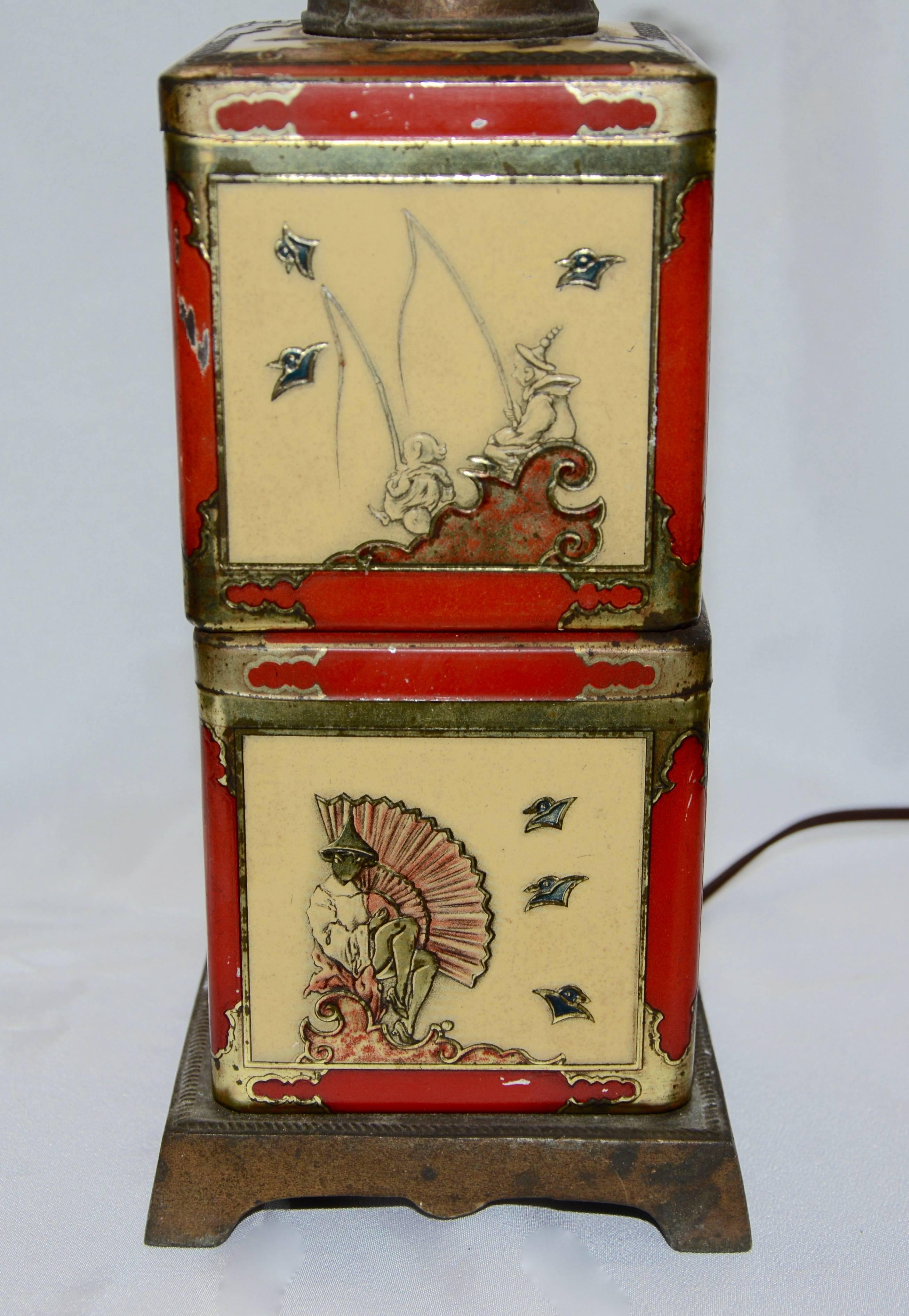 A pair of Asian antique tea tins make up this electric table lamp that was designed in the 1940s. The tins rest on a cast metal base. There are two different scenes on each tin. A cast metal piece with a faux key sits atop the tins and is completed