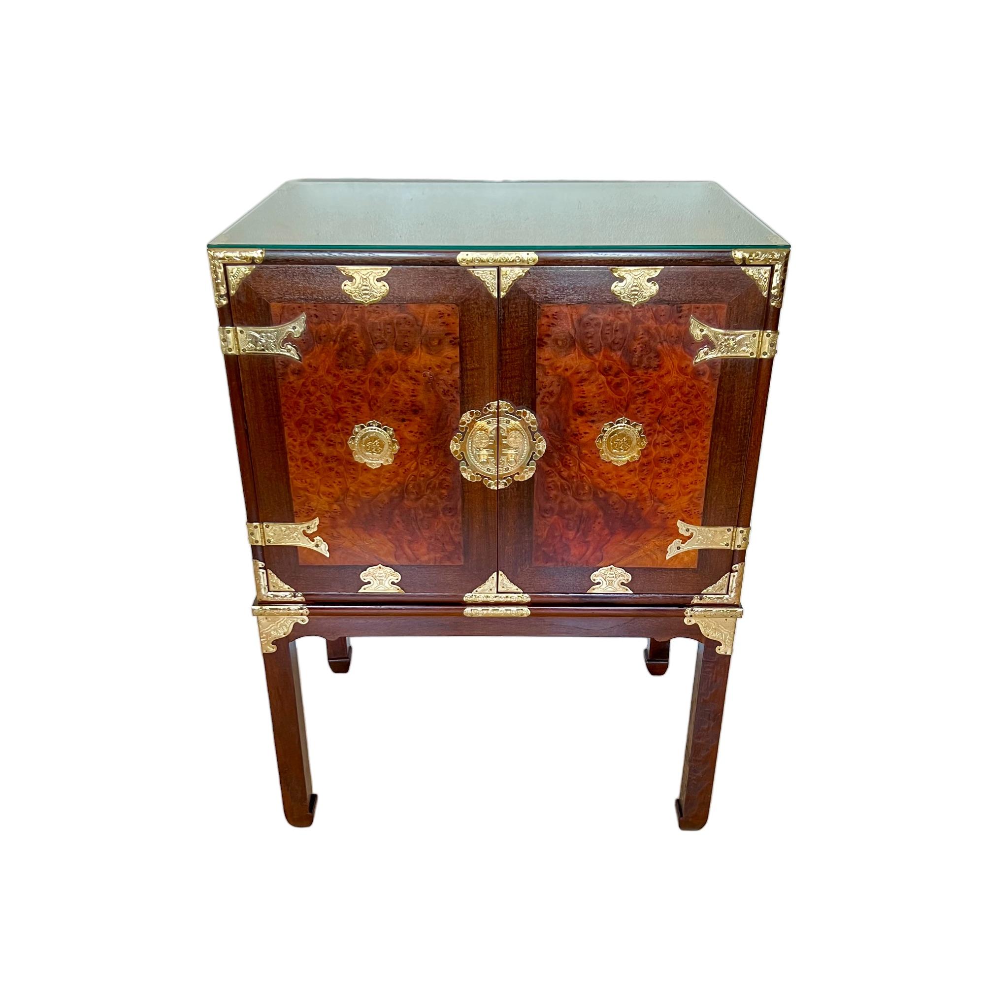 This vintage 1970's Chinese campaign style cabinet on stand is finely crafted of teak and burl elm wood and embellished with etched brass mounts. This chinoiserie storage piece features a spacious two door medallion adorned cabinet with heavy brass