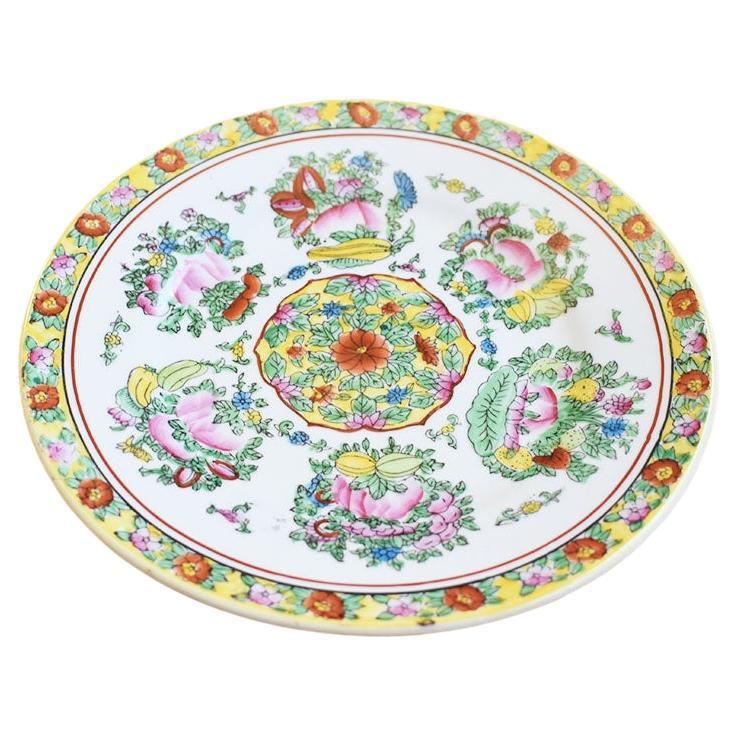 A small ceramic decorative chinoiserie plate. Decorated in yellow around the edges, this plate features chrysanthemums and brightly painted fruits in yellow, red, and pink among lush green leaves. A hanger has been applied to the back to make