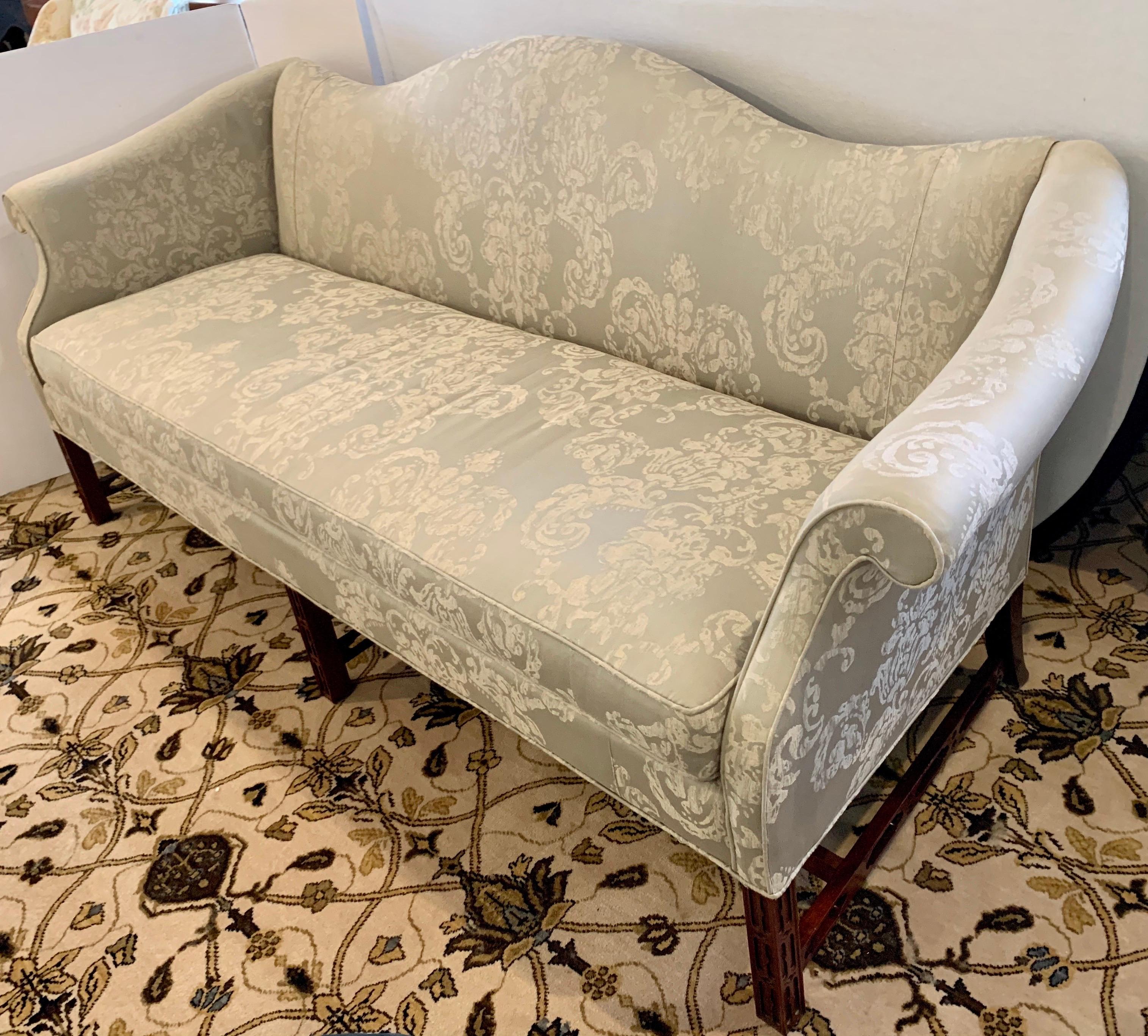Classic and elegant Chinese Chippendale camelback sofa features a shapely mahogany frame with scrolled arms and single thick cushion. Supported by six elegant tapered legs conjoined with pierced fretwork stretchers. Upholstered in a luxurious silver