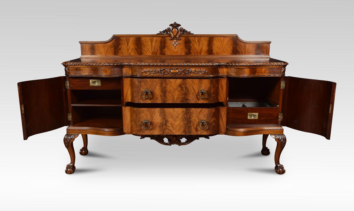 Flame mahogany sideboard. The raised back with central carved motif, above the large breakfront top with gadroon carved edge. The sideboard is fitted with three central draws having tooled brass handles and mahogany draw liners. Flanked by cupboard