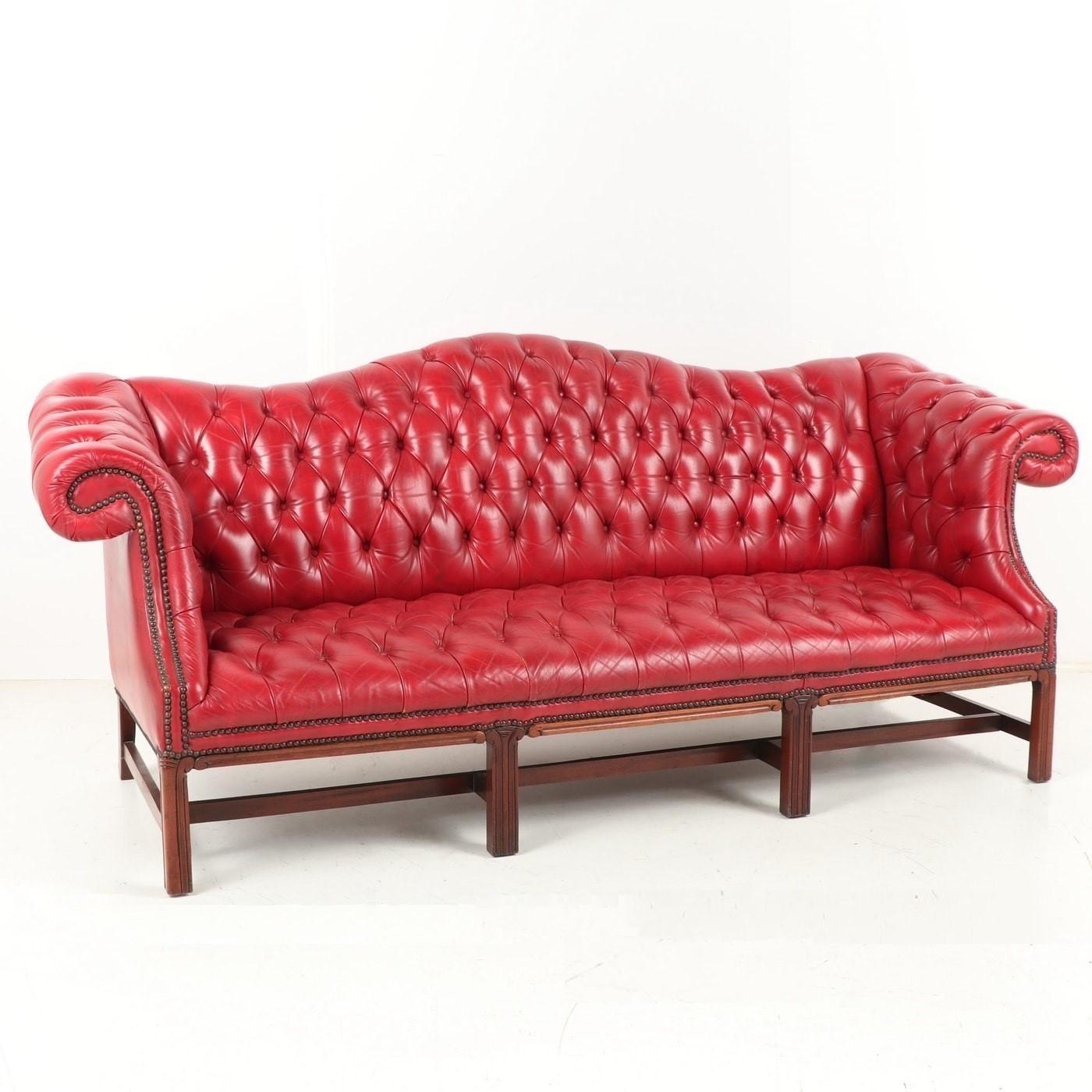 Camel back sofa in the Chippendale style with Marlborough fluted front legs braced with the sabre rear legs through stretchers. The leather is red, supple and deeply buttoned, finished with closely nailed brass studs at the edges. The proportions of