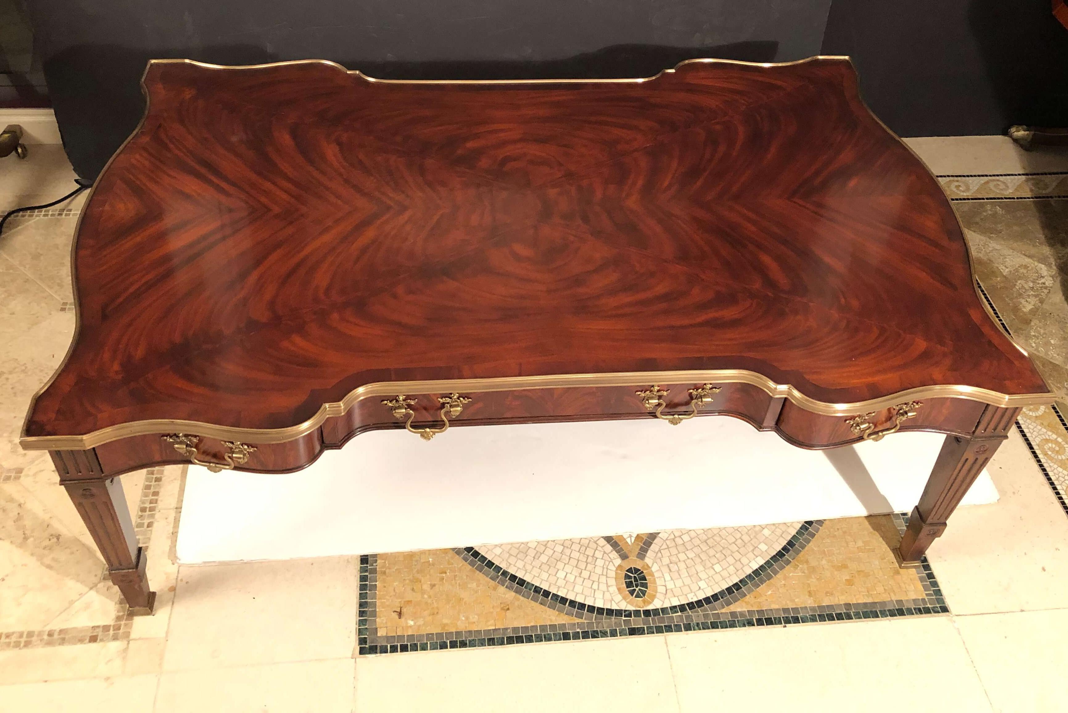 A large Chippendale style flame mahogany veneer and solid cocktail table, the serpentine brass bound top above two drawers on each side centered by a leather inset slide drawer with a concealed drawer, all with brass Rocaille handles, on square