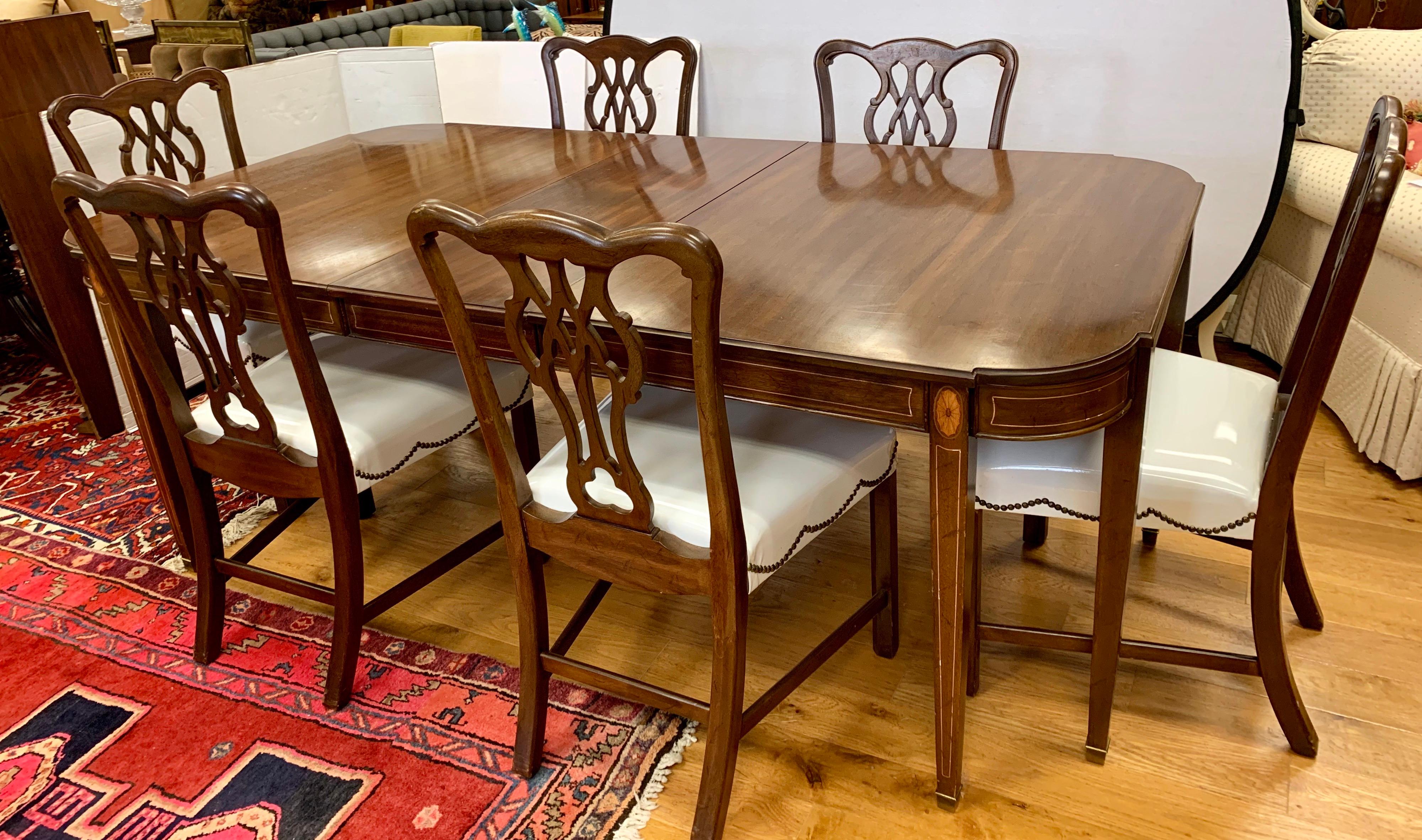 Classic Chippendale style dining set includes a mahogany extension table with oval and line inlay. It rests on eight square tapered legs with brass caps and has rounded corners. The two leaves measure 16” wide. Table is shown with one leaf