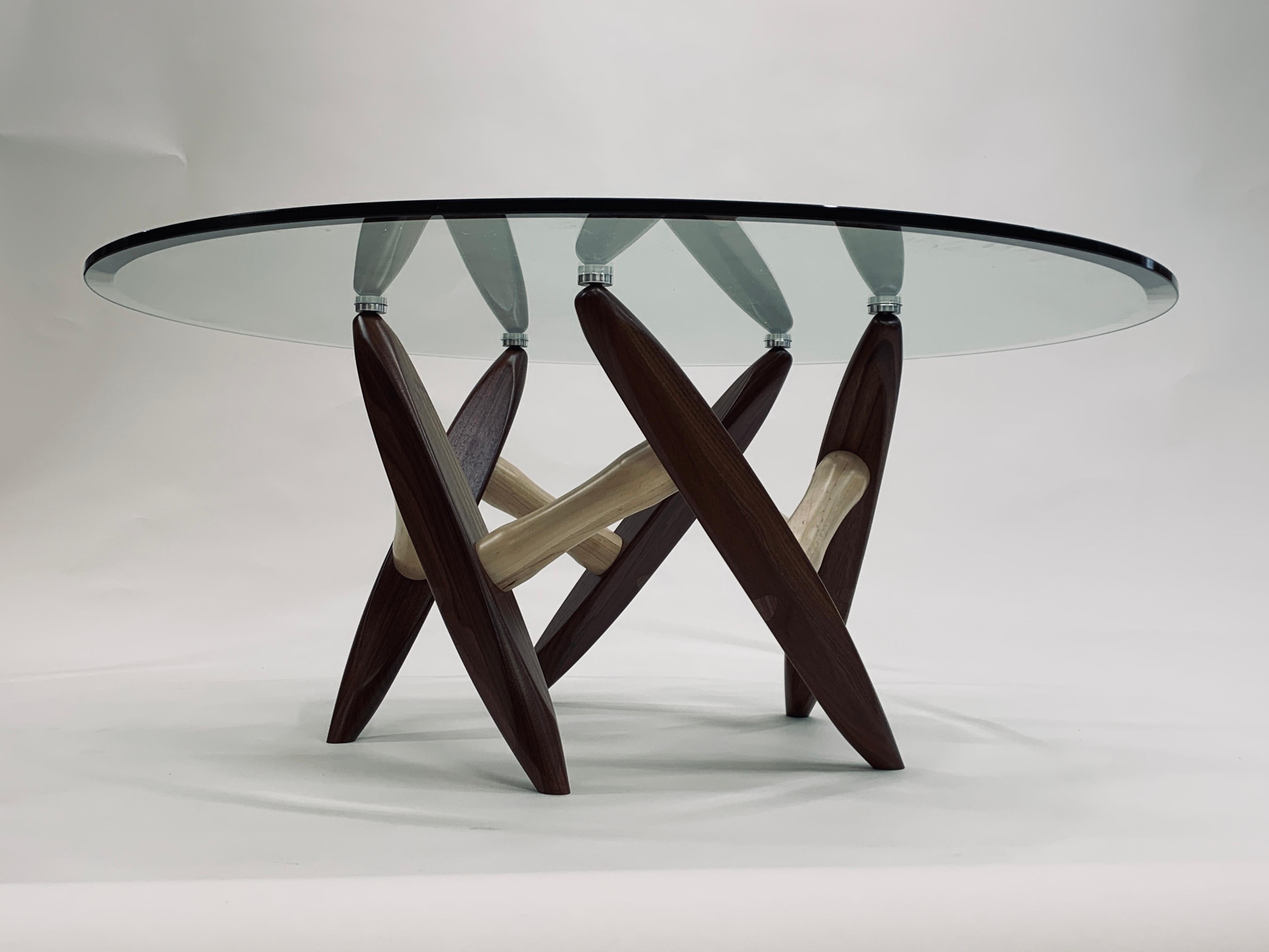 El tablero de cristal de la mesa de centro Chiral K531-40 descansa sobre una escultural base de madera maciza que, a su vez, es un ejercicio de simetría radial.  Las cinco patas de nogal, cada una con sección transversal triangular, se inclinan unas