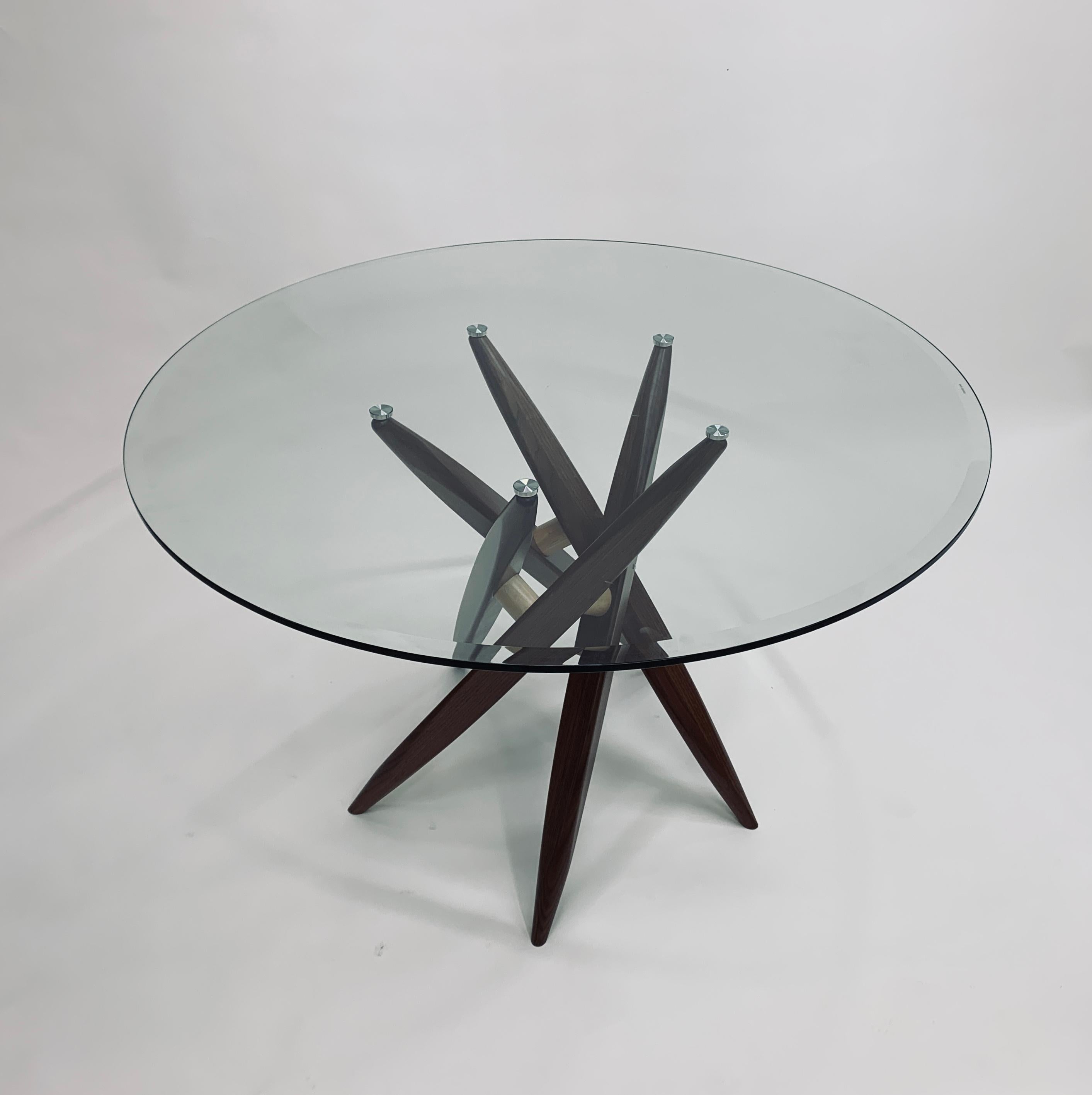 Il piano in vetro del tavolo Chiral T531-44 poggia su una base scultorea in legno massiccio che è essa stessa un esercizio di simmetria radiale.  Le cinque gambe in noce, ognuna con una sezione trasversale triangolare, si appoggiano e si superano