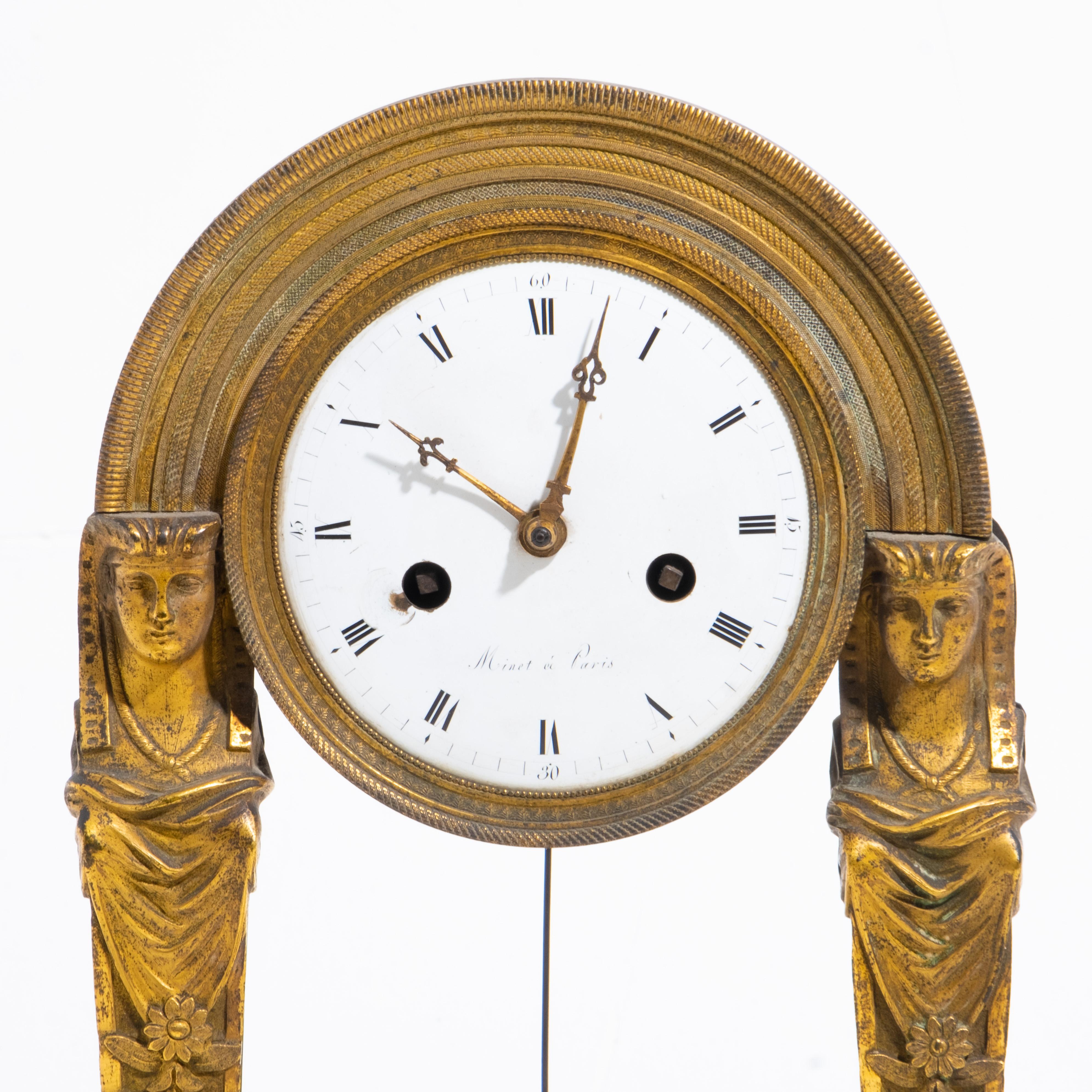 Diese Uhr aus der Restaurationszeit ist aus gemeißelter und vergoldeter Bronze gefertigt und besitzt ein weiß emailliertes Zifferblatt, das von Minet à Paris signiert ist. Die Uhr wird von zwei Begriffen im ägyptischen Stil getragen, die die