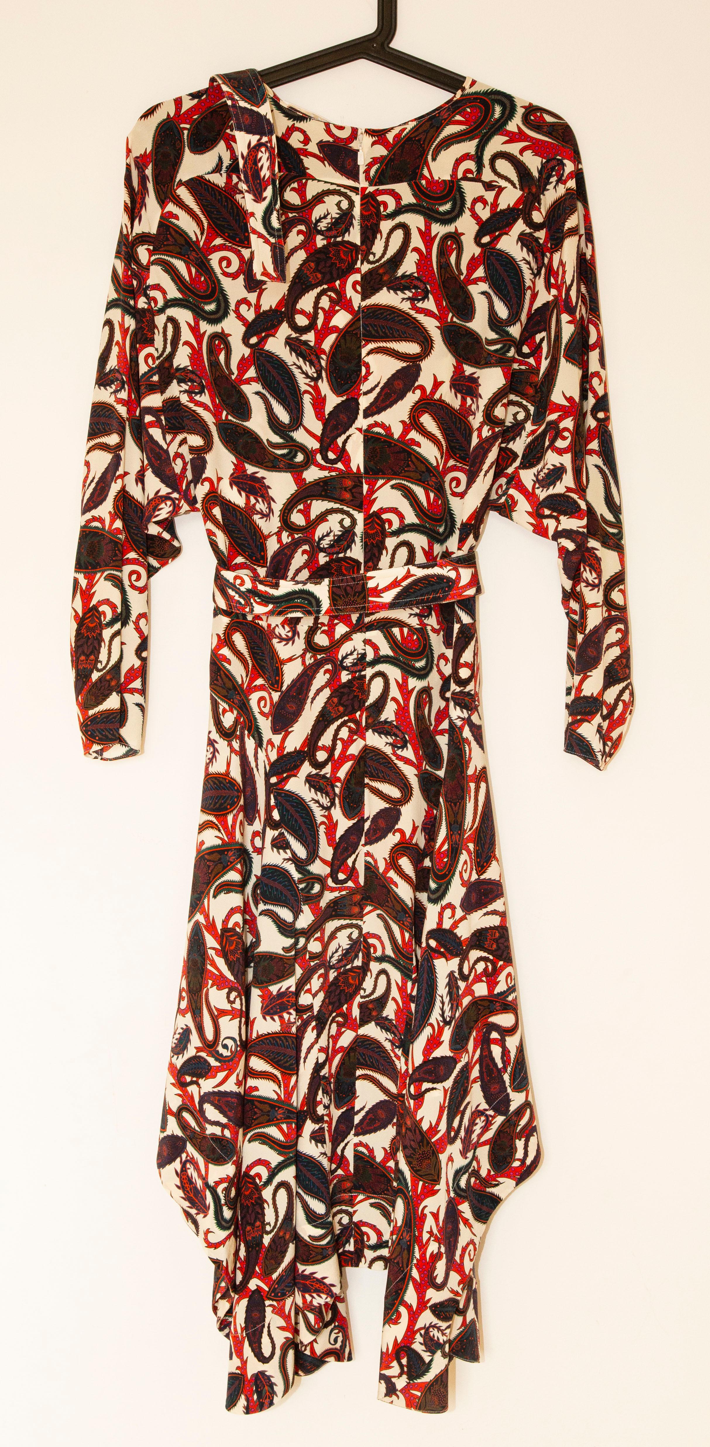 A Chloe silk mid length dress with blue, red, green, pink paisley print. The dress features  asymmetric bottom, long sleeves, and belt with a silver toned buckle. The condition is very good.
