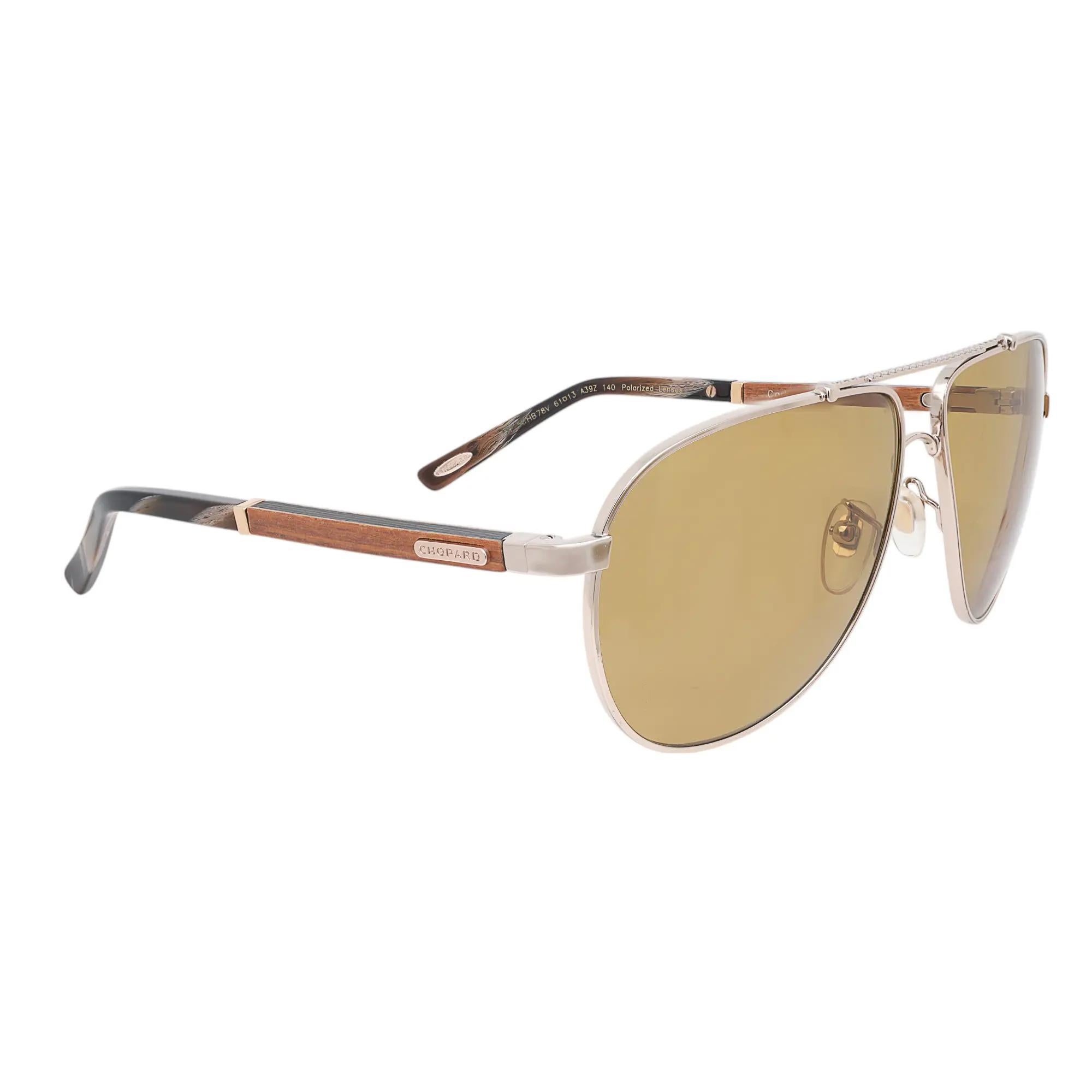 Presenting the luxury unisex Chopard Sunglasses 95221-0417, ideal for street fashion. These sunglasses feature a shiny light gold/ebony metal frame with wood-carbon fiber color, ocher polarized A/R lenses. The arms are adorned with the Chopard logo.