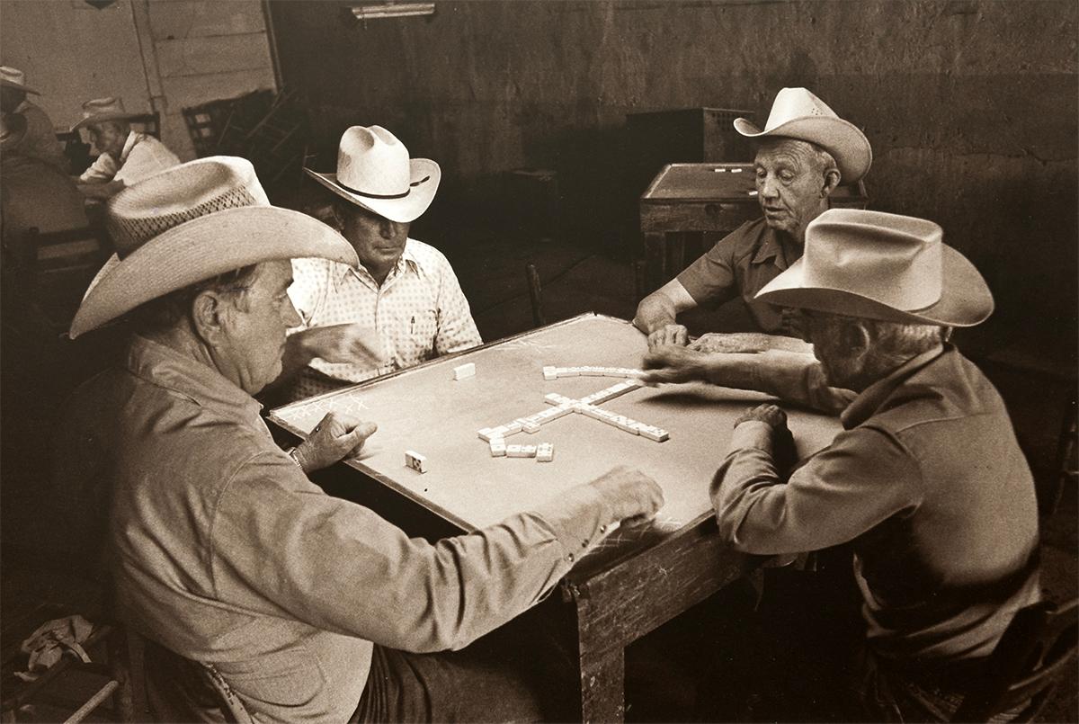Domino Players, Lawn, TX by Chris Regas depicts four men wearing cowboy hats sitting at a table, engaged in a game of dominos. 

This silver gelatin print measures 9.25 x 15.5 inches, and is edition 6/10. 
This photograph is signed in pencil on