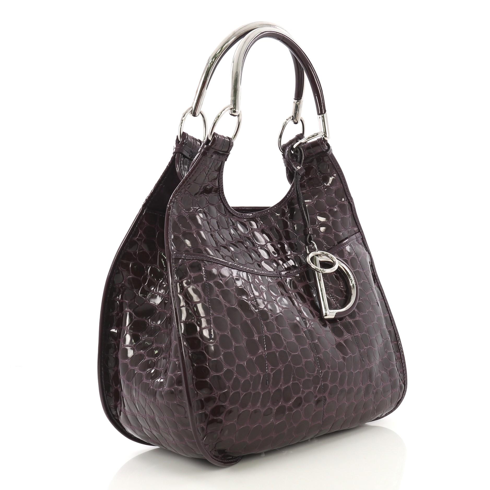 This Christian Dior 61 Shoulder Bag Crocodile Embossed Patent Medium, crafted in purple crocodile embossed patent leather, features dual metal handles, exterior front slip pockets, Dior charms, protective based studs and silver-tone hardware. It