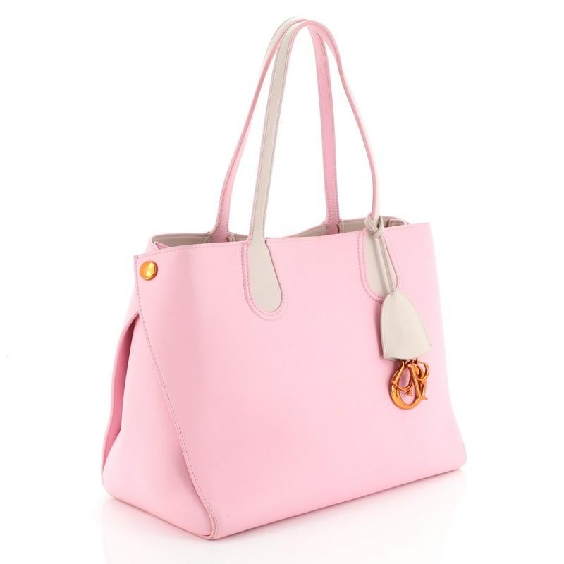 This Christian Dior Addict Shopping Tote Leather Small, crafted in pink leather, features dual slim leather handles, side snap buttons, Dior charms, and bronze and silver-tone hardware. Its magnetic snap closure opens to a neutral leather interior.