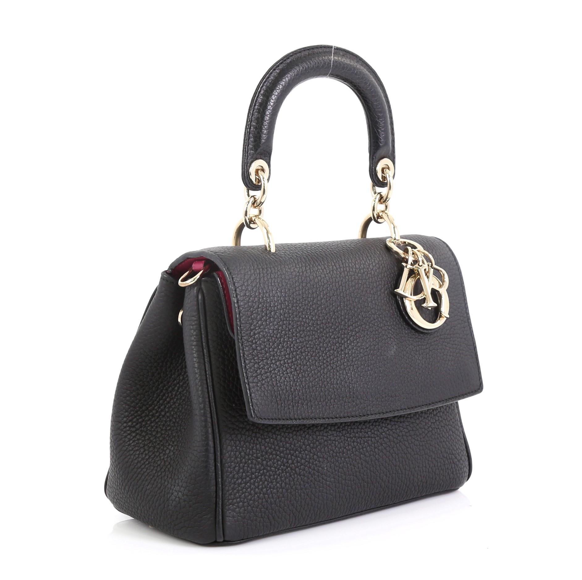 This Christian Dior Be Dior Bag Pebbled Leather Small, crafted from black pebbled leather, features leather handle with Dior charms, side snap buttons, exterior back pocket, protective base studs and gold-tone hardware. Its flap opens to a pink
